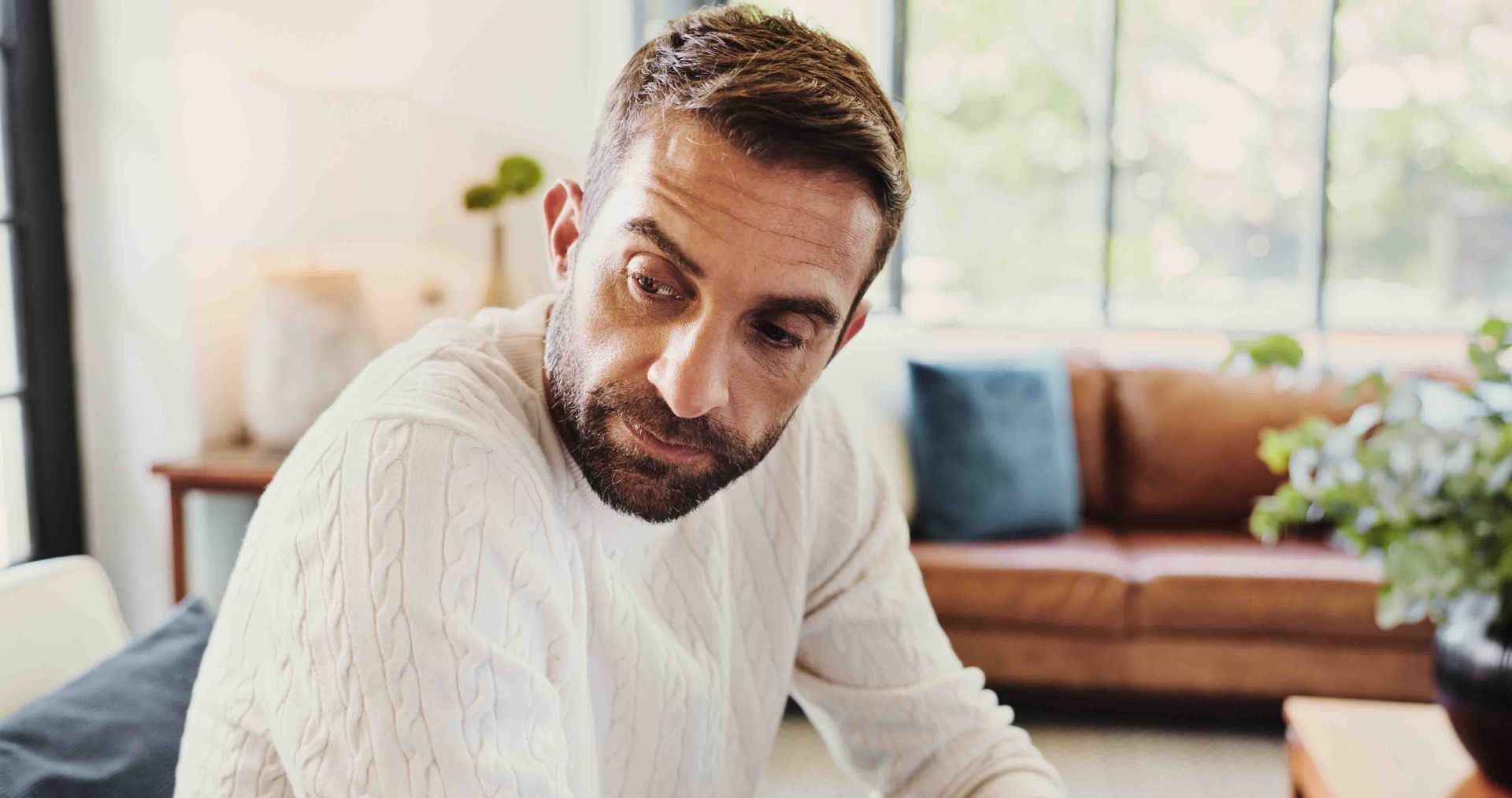 Man with a beard in a cream sweater, looking down with a focused expression. Interior shot with brown couch.