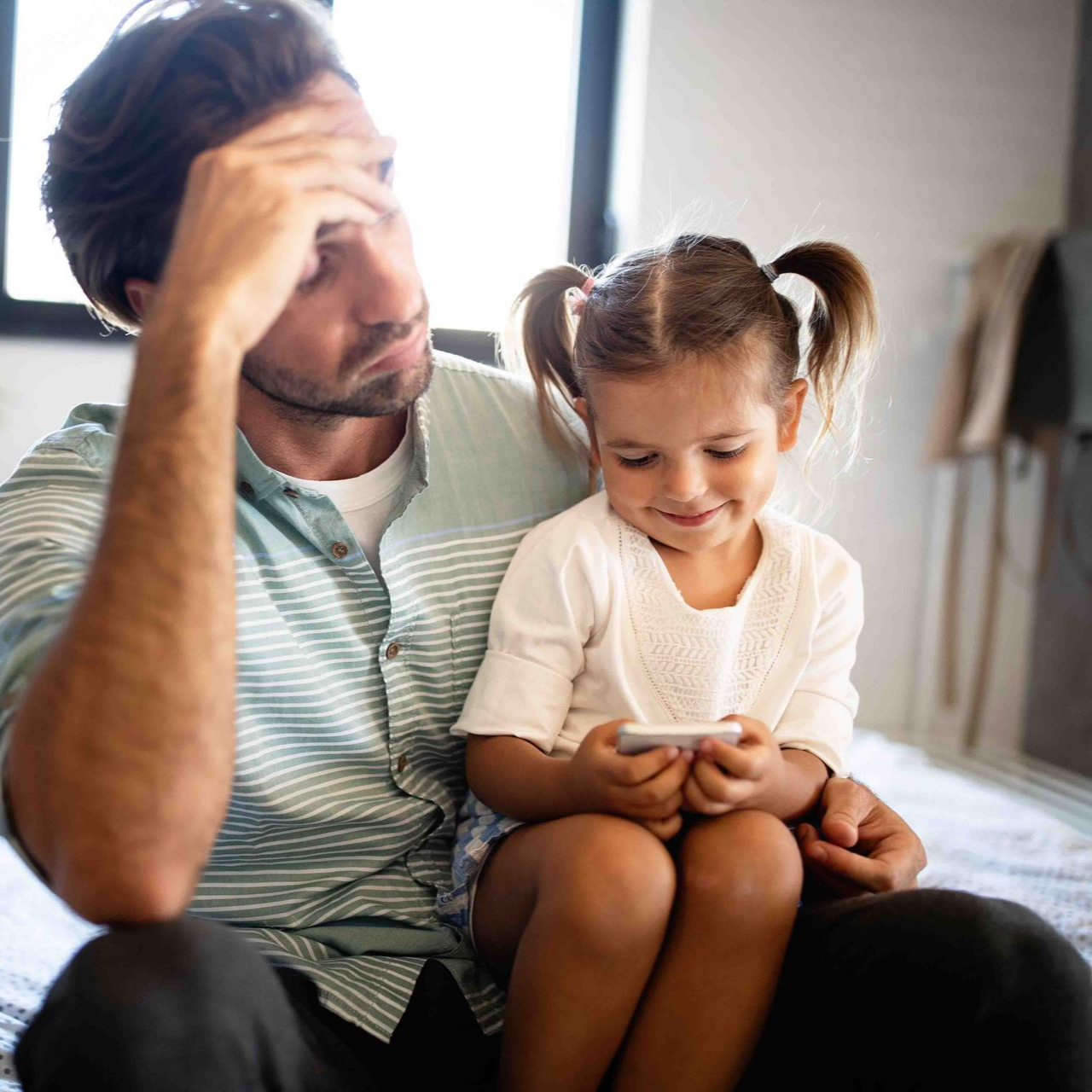 Man with hand on forehead, looking stressed, sitting next to a child focused on a phone.