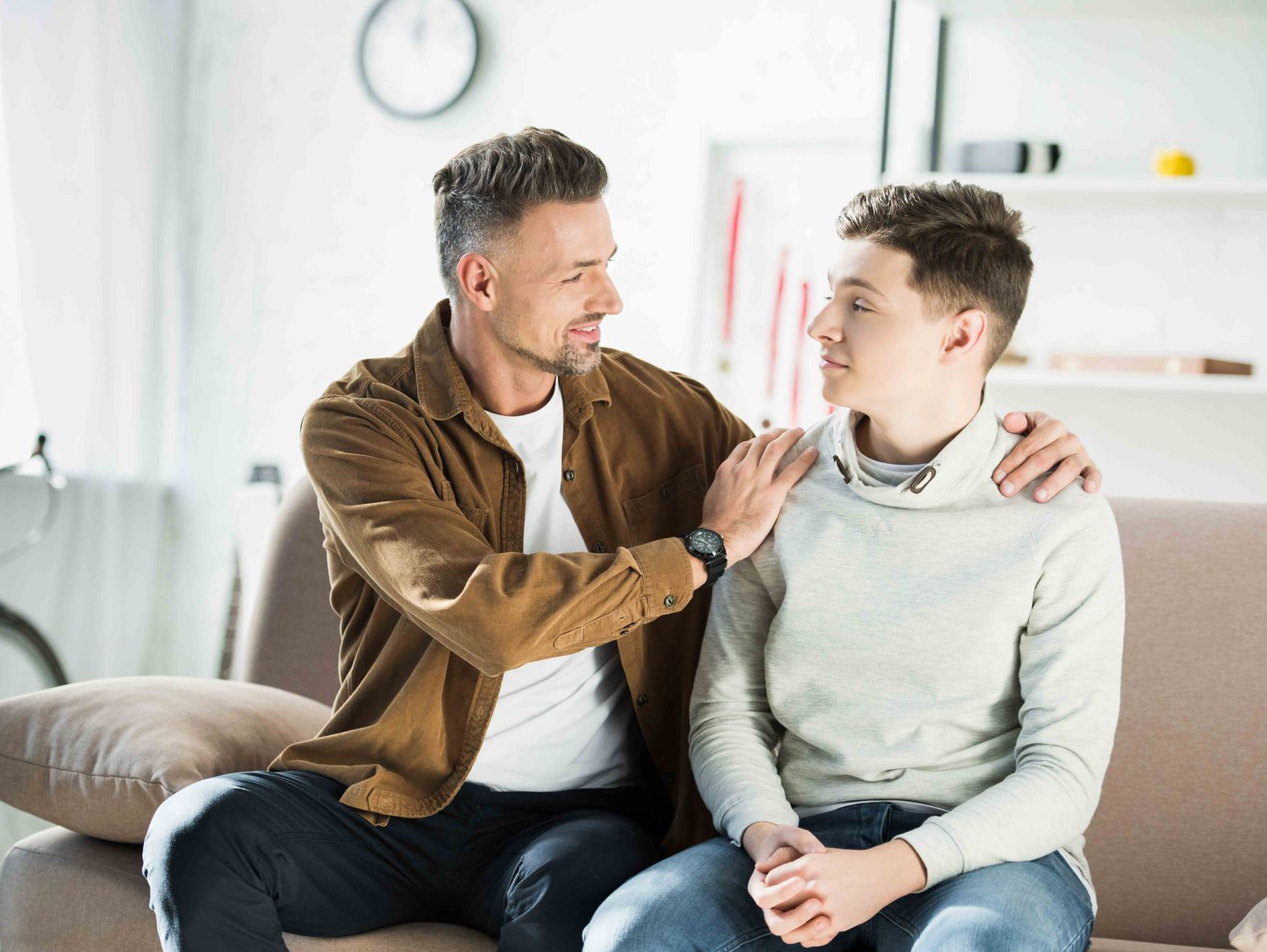 Man with hand on son's shoulder, smiling at him while sitting on a couch. Living room setting.