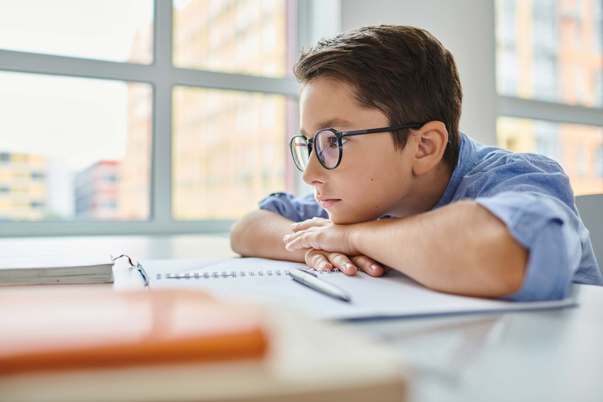 Boy wearing glasses, leaning on arms at a desk, looking out a window. Notebook and pen in front.