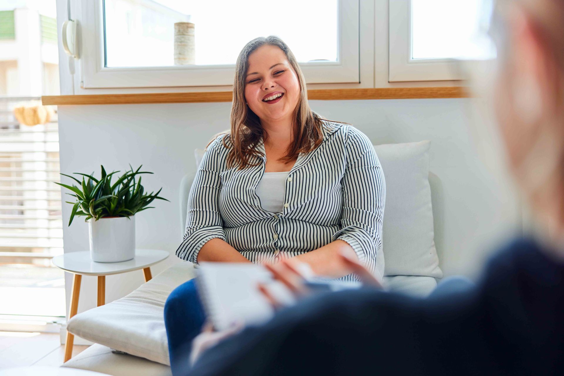 Woman smiling, seated indoors, facing someone holding a notepad. White and black patterned shirt. Plant on side table.