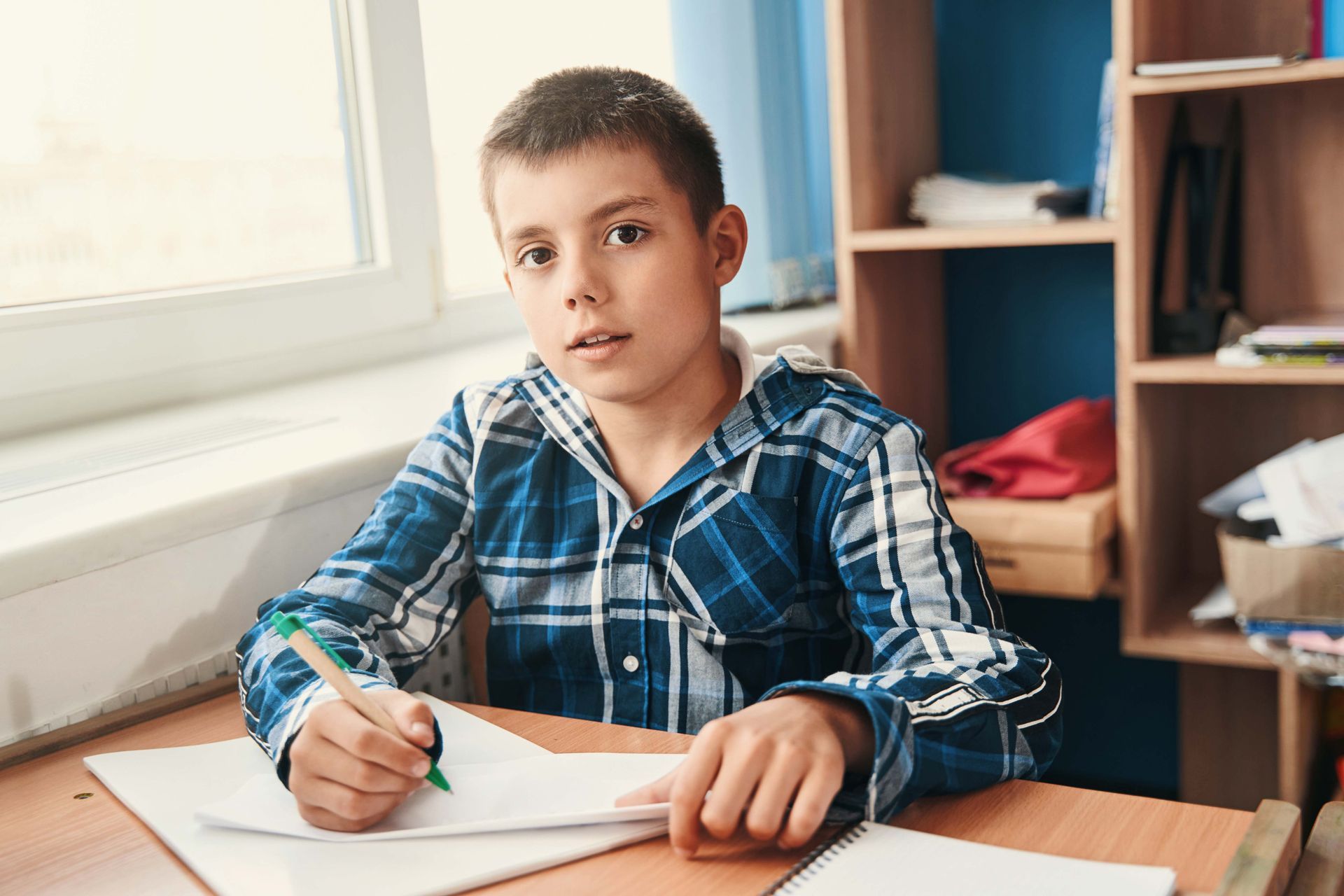 Boy writing at a desk with a plaid shirt, looking at the viewer. A window and shelves are in the background.