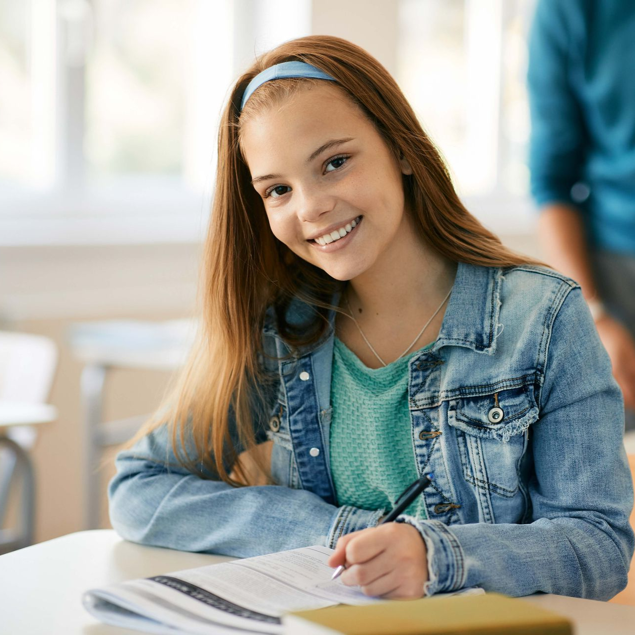 Girl with long brown hair, wearing a denim jacket, smiles while writing at a desk in a classroom.