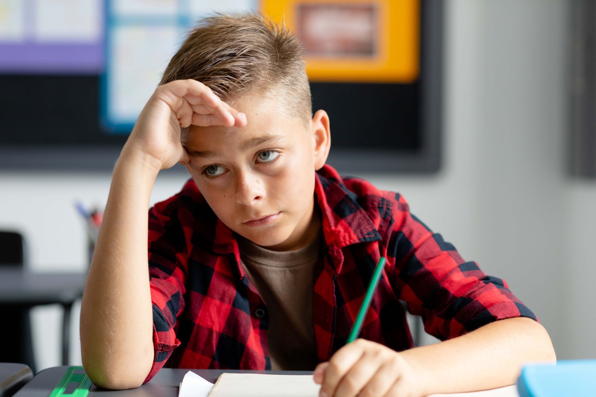 Boy in red plaid shirt looking frustrated, hand on forehead, pencil in hand, at a desk in a classroom.