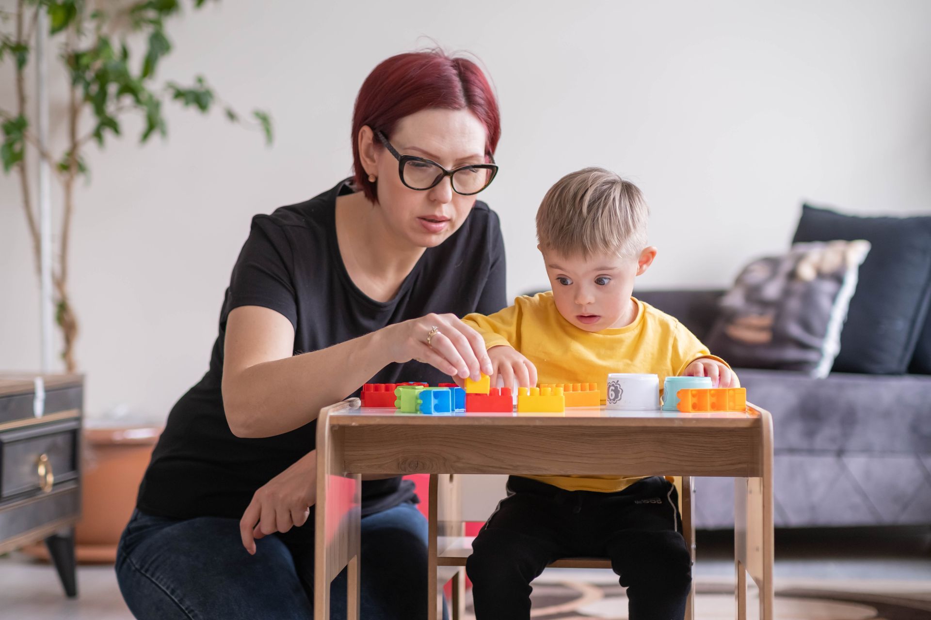 Woman and child with Down syndrome playing with colorful blocks at a small table indoors.