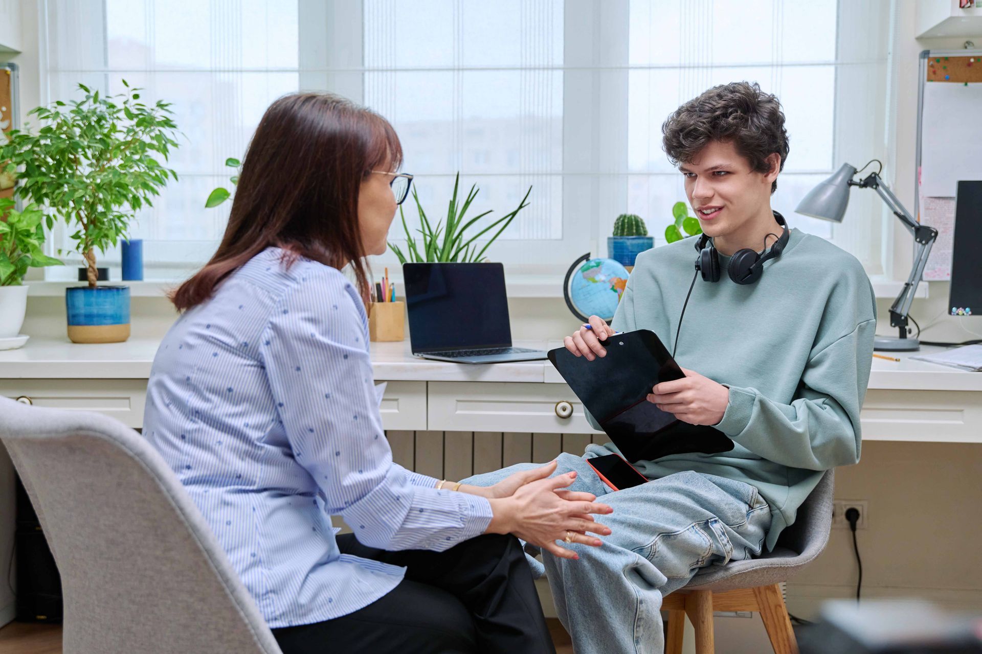 Woman and teenager in conversation at a desk, likely therapy session. Both seated, woman facing teen, holding hands.