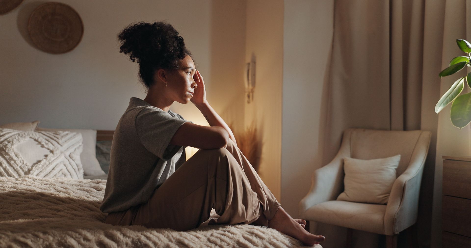 Woman sitting on bed, looking thoughtful, in a warmly lit bedroom.