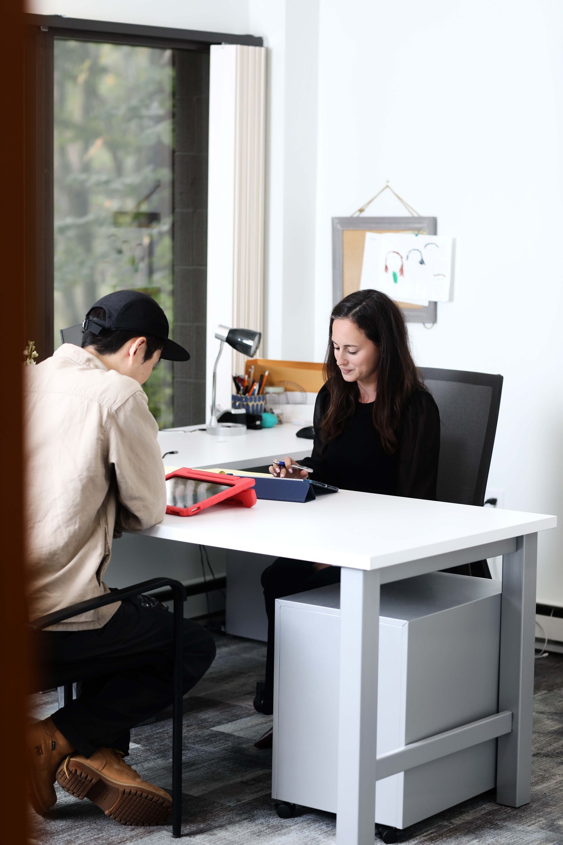 Person wearing a cap and woman seated at a desk, looking at tablets. Office setting with a window.
