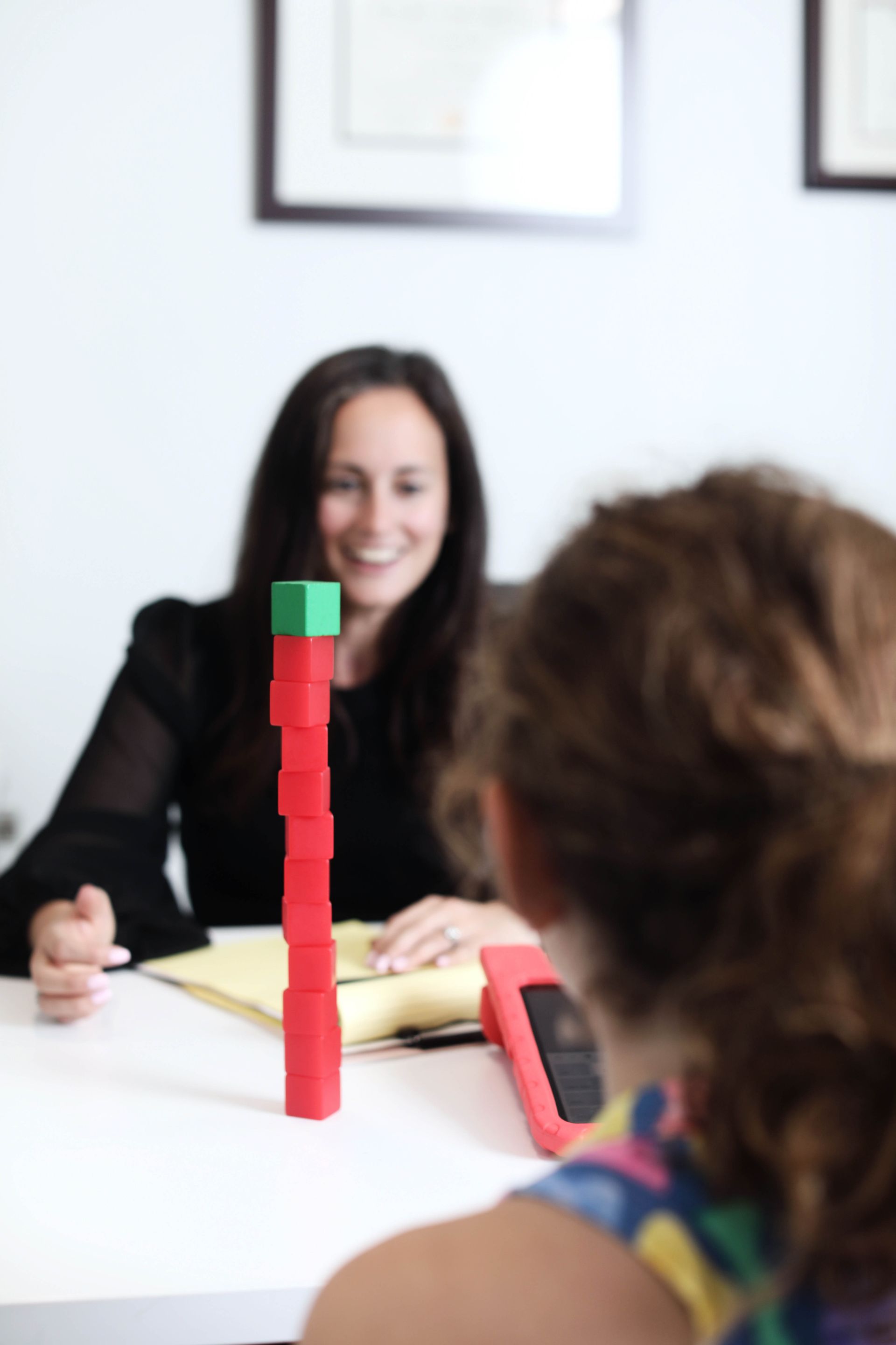 Woman smiles at a child, both seated at a white table. A tall stack of red blocks with a green top sits between them.