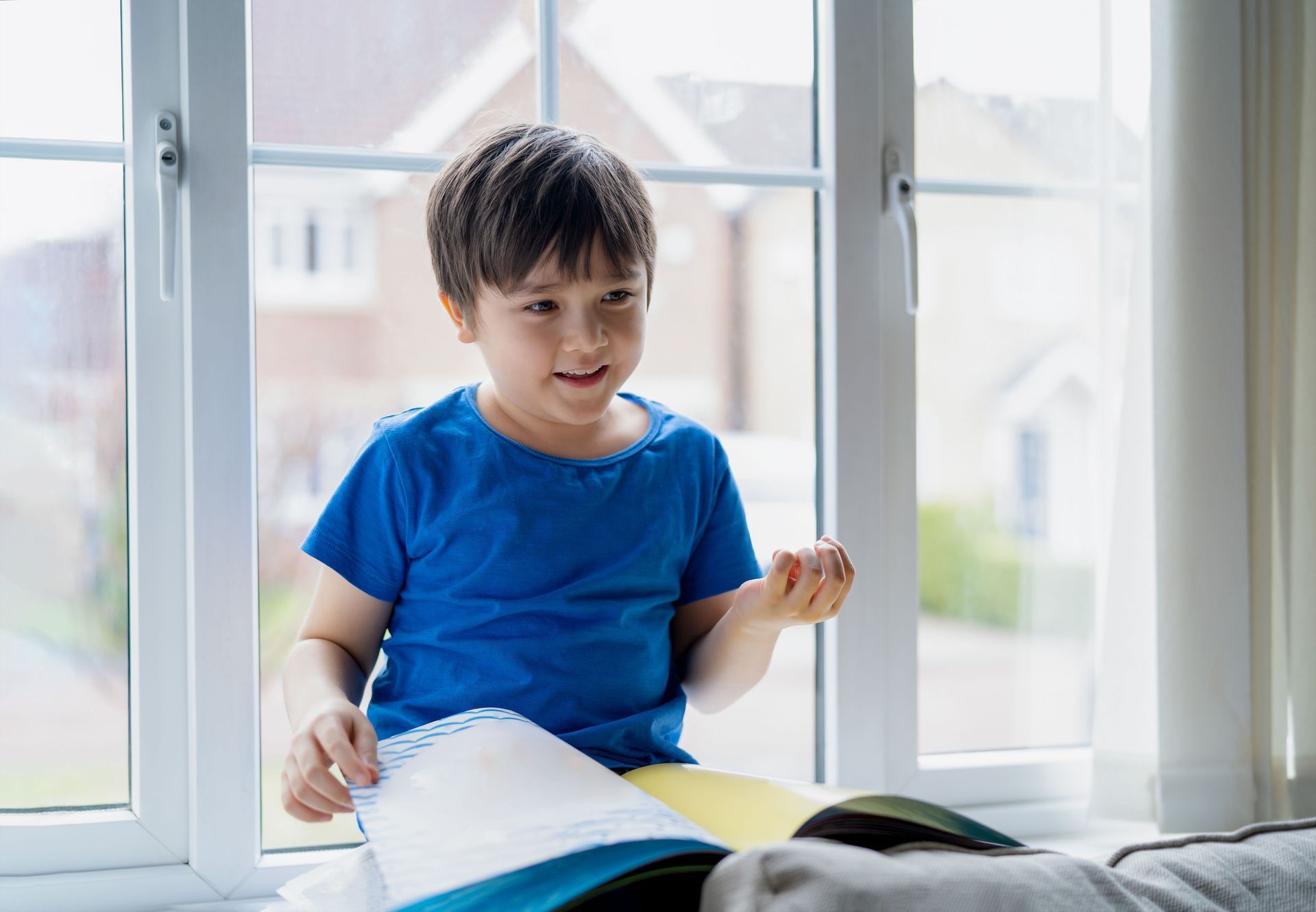 Young child in a blue shirt looks at a book, sitting on a windowsill, smiles.
