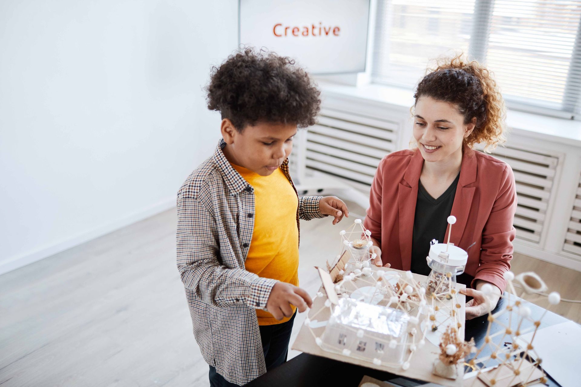 A child and adult build a model structure together at a table in a brightly lit room.