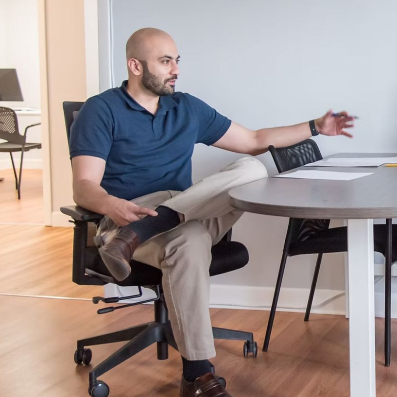Man in blue shirt and khaki pants gestures while seated at a round table, leg crossed.