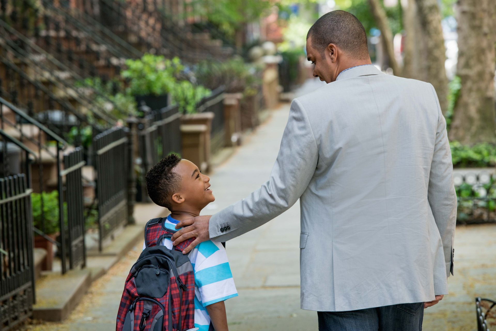 Man with hand on boy’s shoulder; they face each other on a sidewalk, backpacks visible.