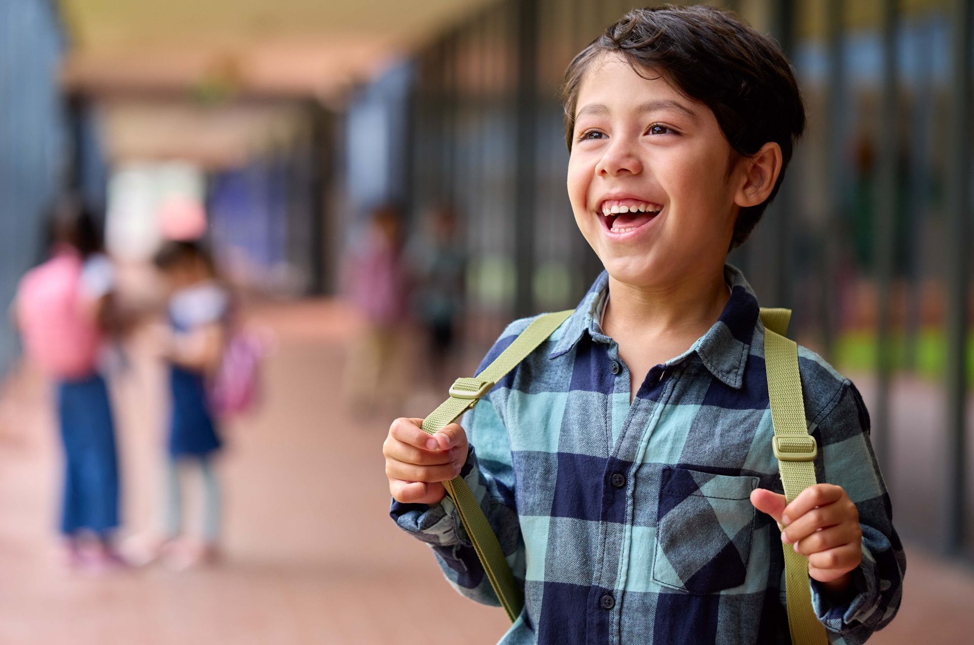 Boy with a backpack smiles broadly in a school hallway.