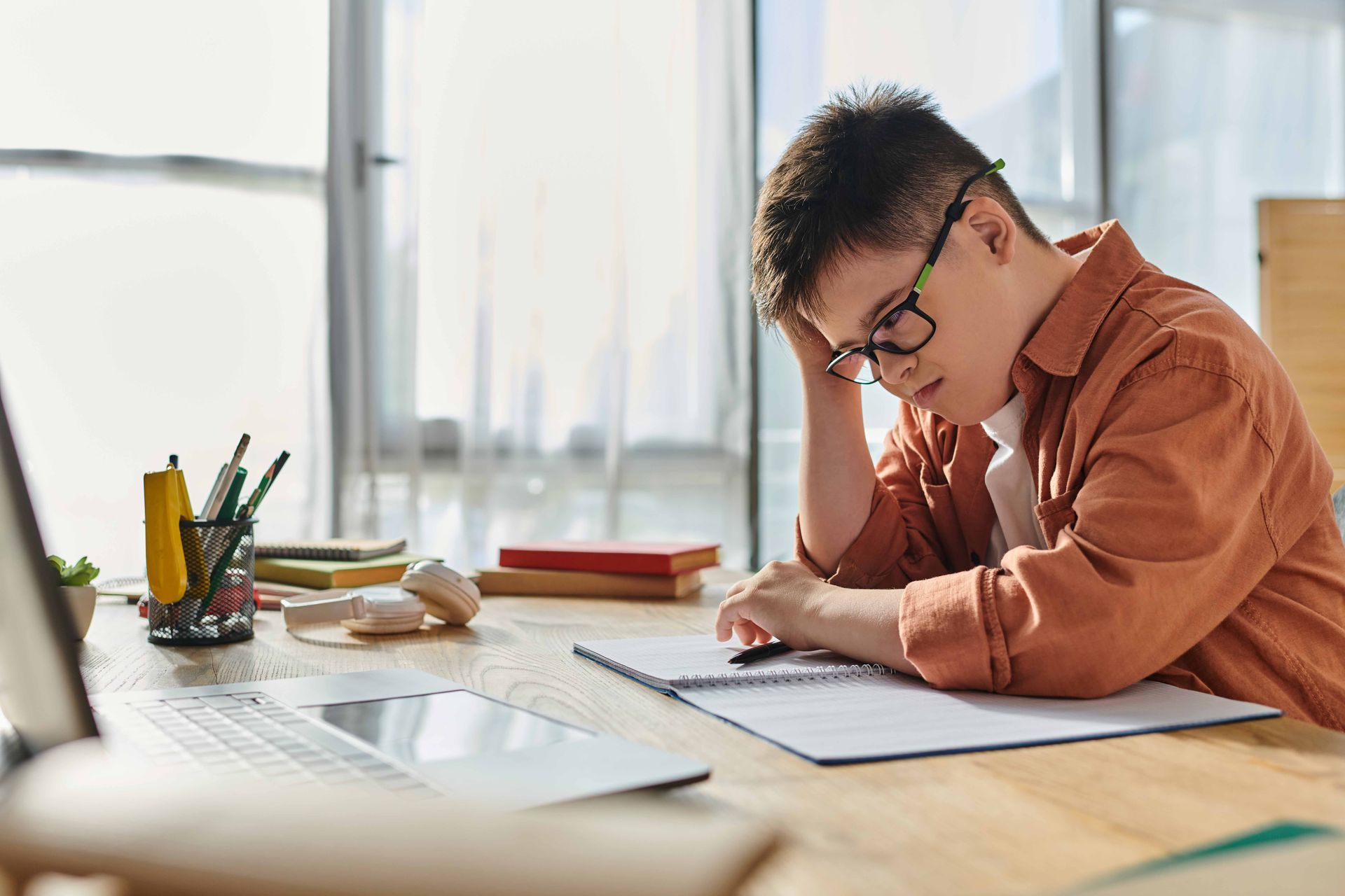 Person with glasses and orange shirt studies at a desk with laptop, notebook, and pencils, looking down.
