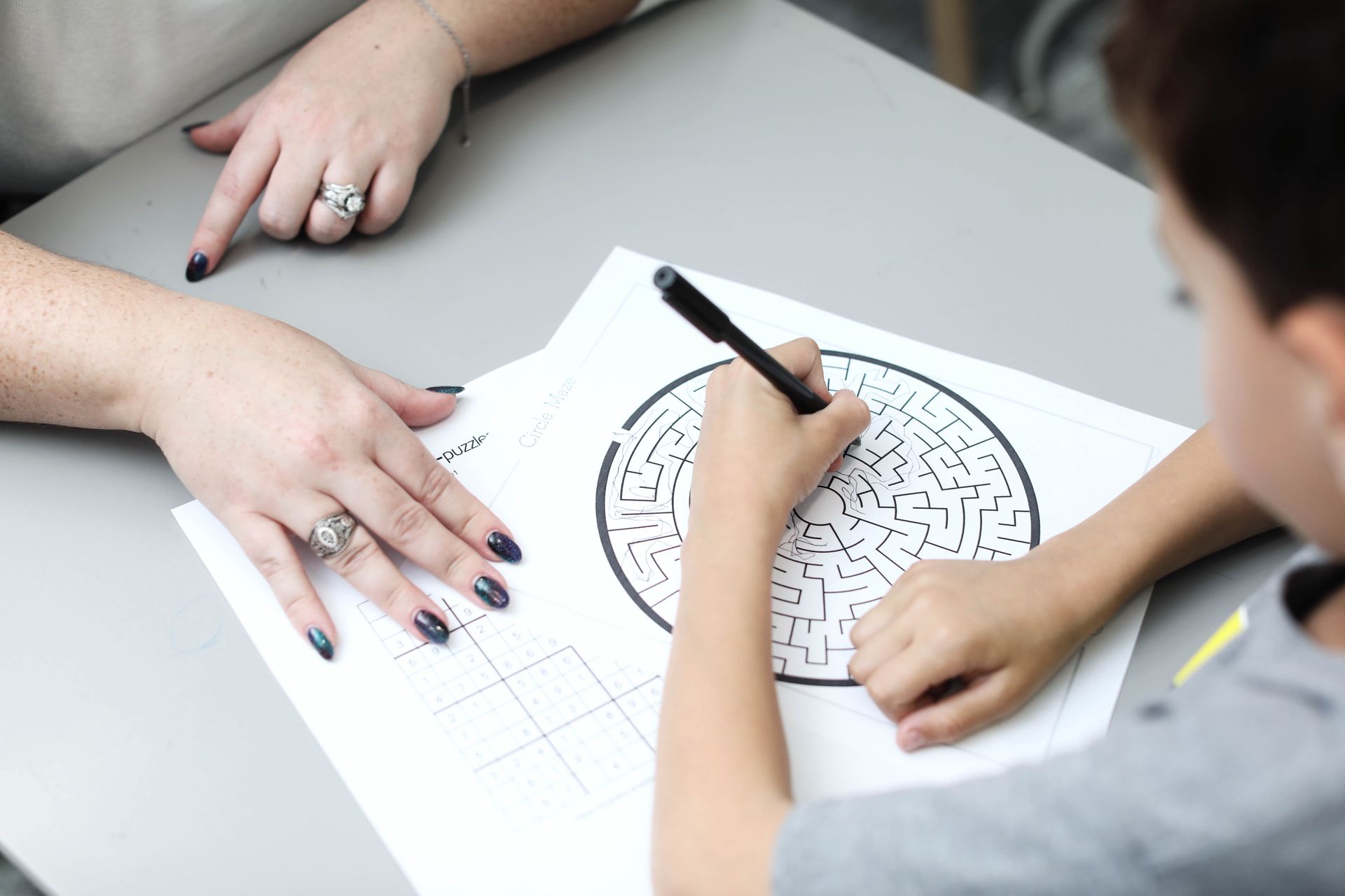 Person assists a child with a maze puzzle. Both are at a table; the person points.