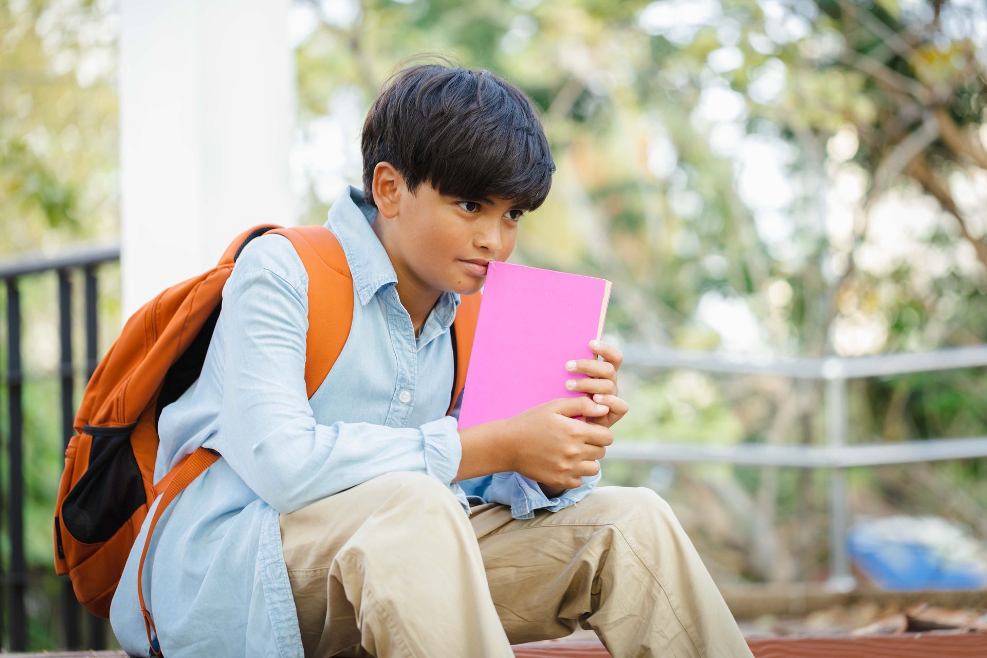 Boy with backpack sits holding a pink book, looking thoughtful, outside.