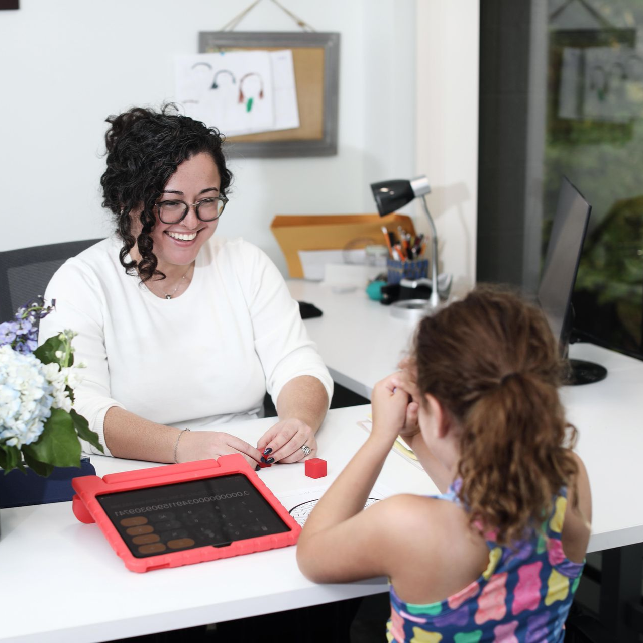 Woman smiling, assisting child with a device at a desk. Flowers and a computer visible.