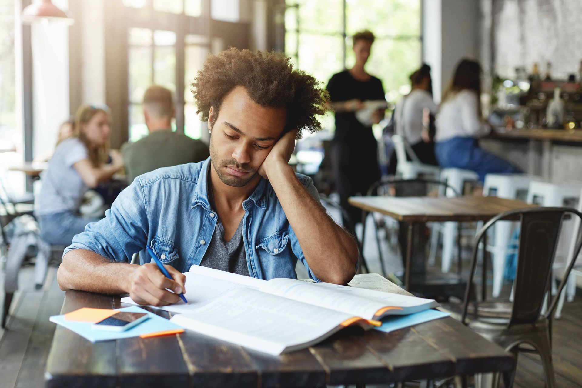 Man leaning on his hand, studying at a cafe table with open books.