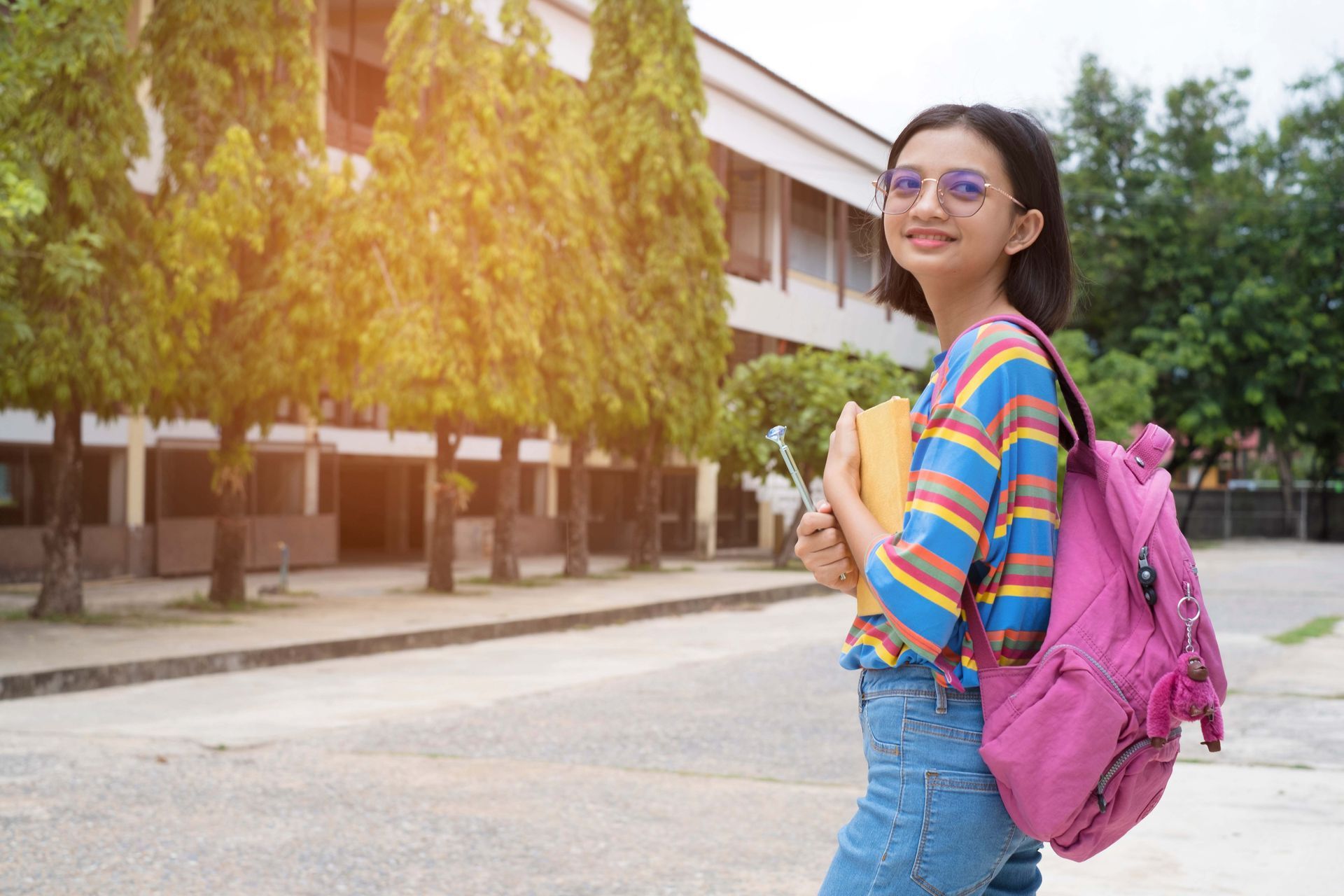 Smiling student with backpack and books stands in front of a school building.