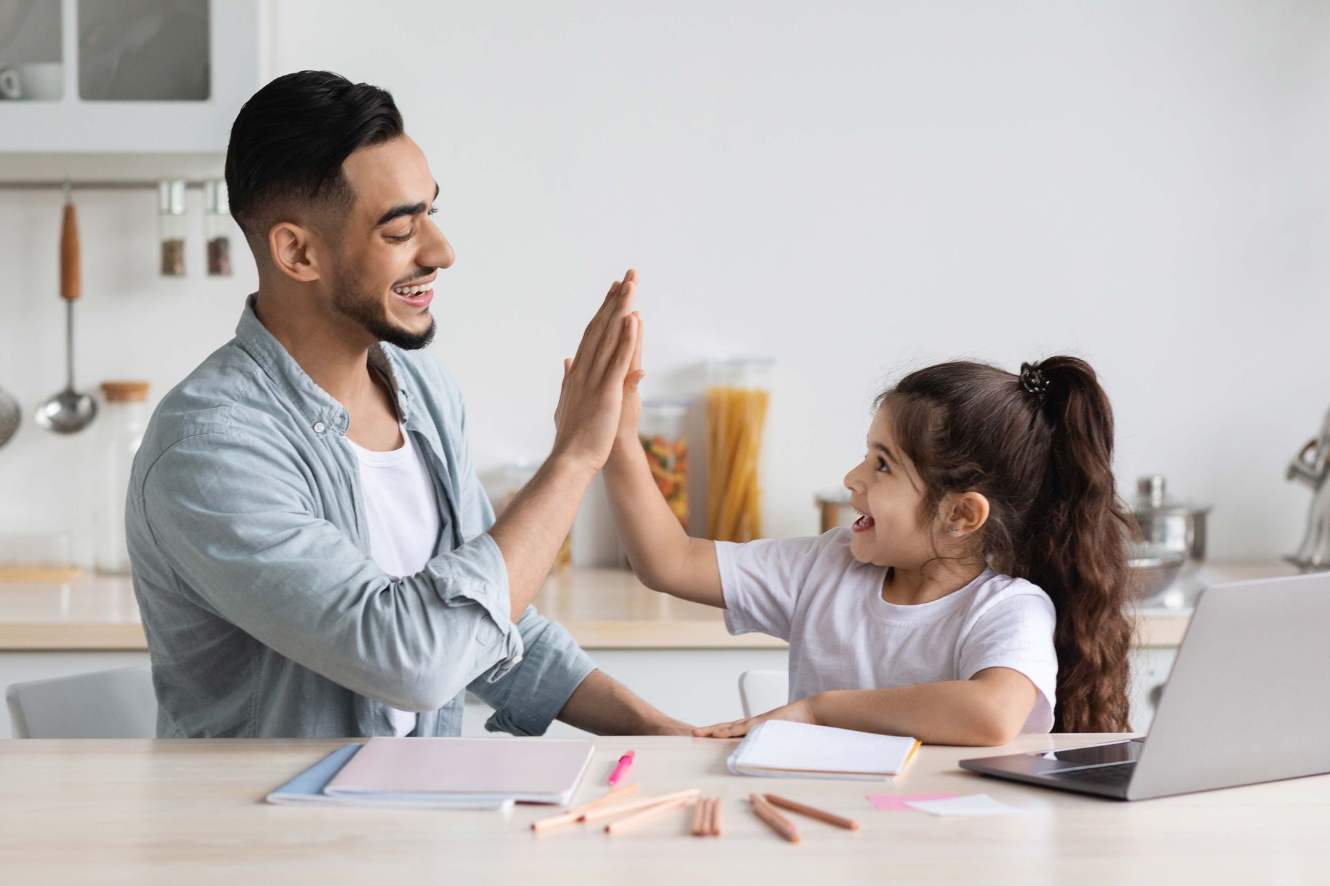 Man and child high-fiving at a table in a kitchen; laptop, paper, and colored pencils present.