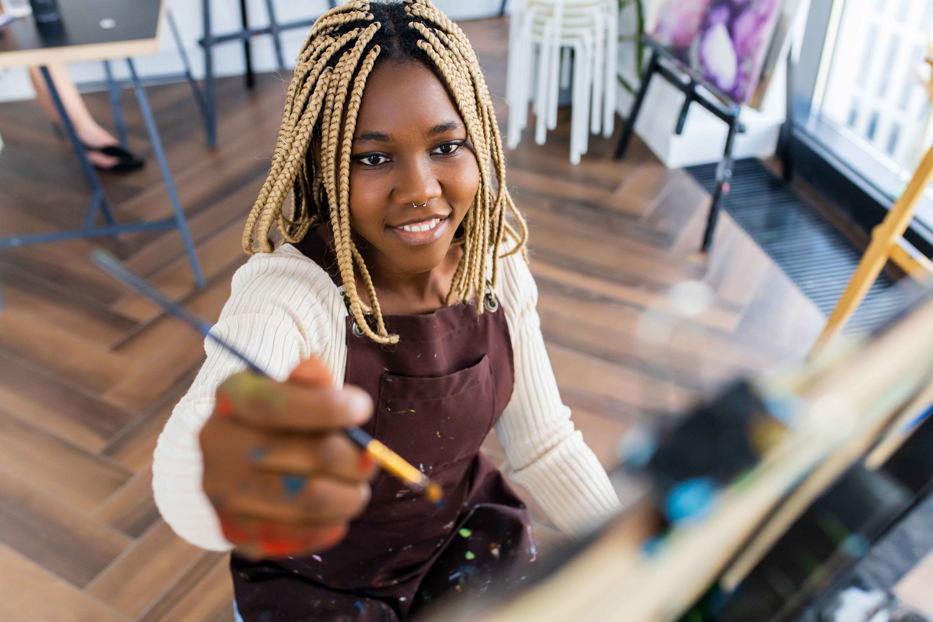 Woman with blonde braids paints on canvas, wearing an apron, in a bright art studio.