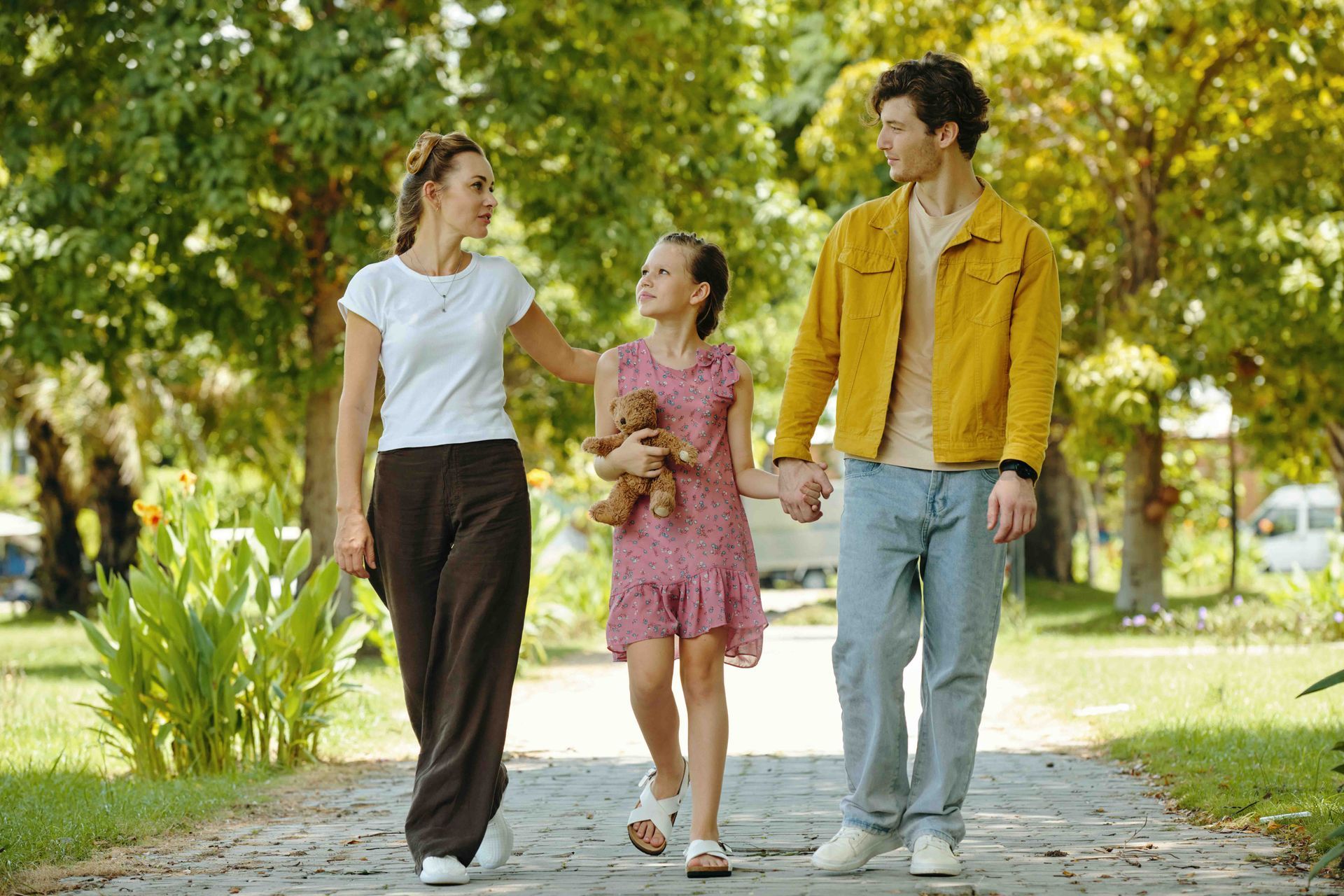 Family of three walking outdoors, holding hands. The child carries a toy. Sunny day, park setting.