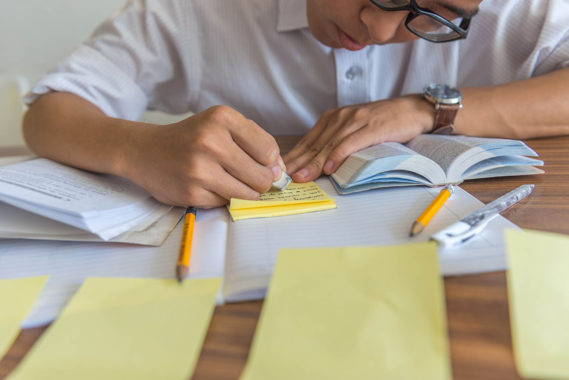 Person wearing glasses erases notes on a sticky pad at a desk with books and pencils.