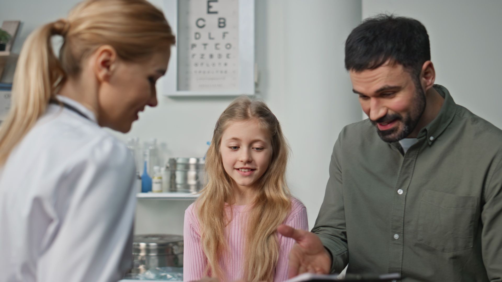 Doctor, girl, and man in an exam room; the man points to a document; eye chart visible.