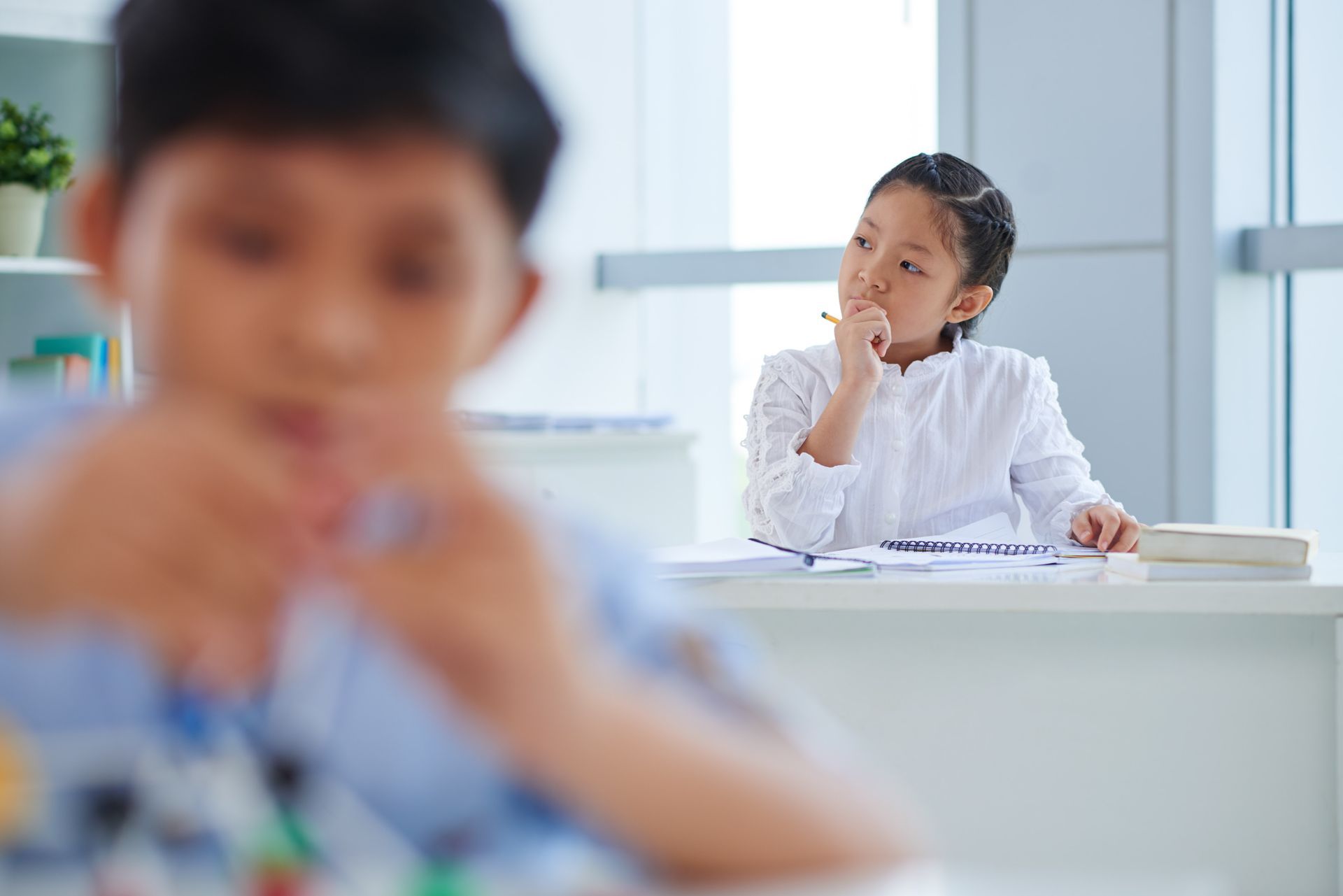 Girl thoughtfully considers something at her desk while boy is blurred in the foreground.