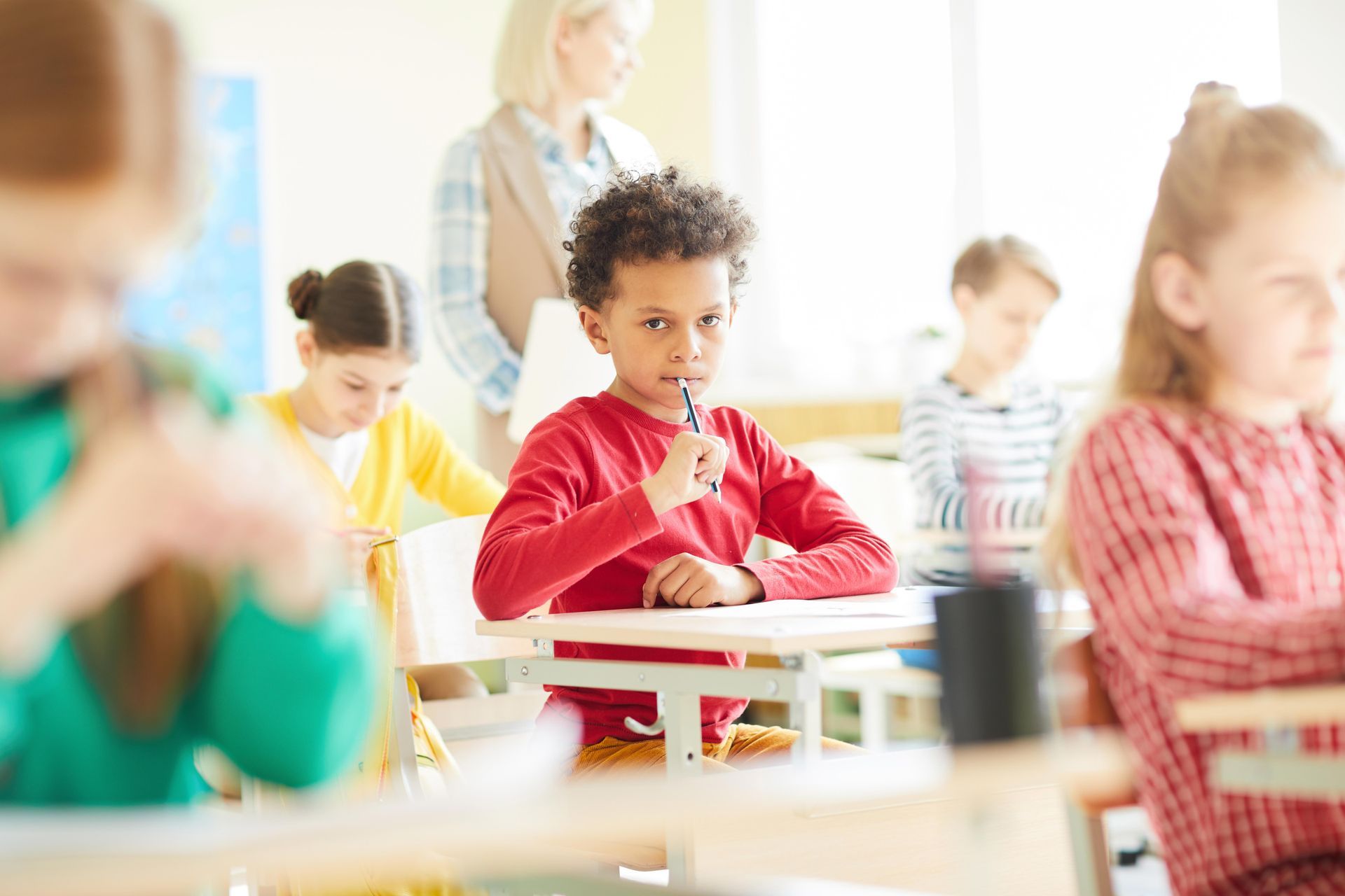 Boy with curly hair in red shirt, thoughtfully holding a pen, sitting at a desk in a classroom.