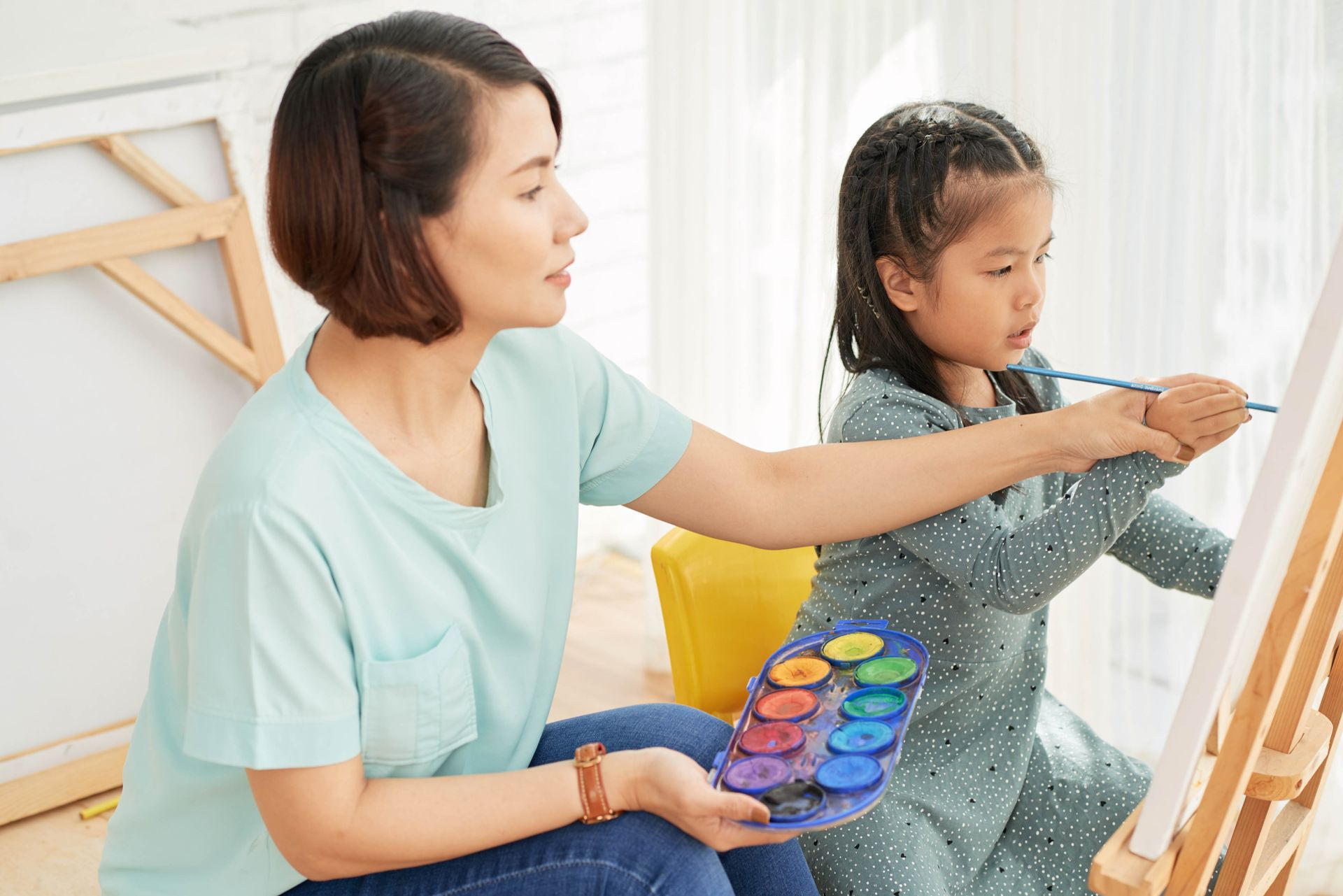 Woman helps child paint with watercolors on easel.