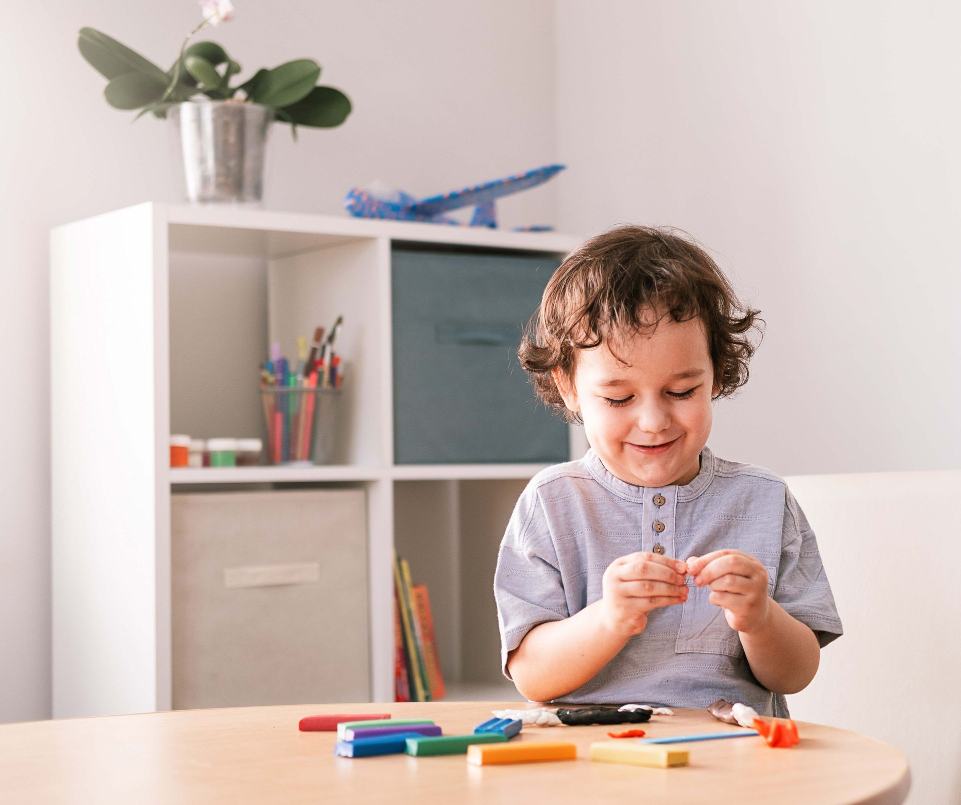 Boy smiles while playing with clay at a table, a shelf with art supplies is behind him.