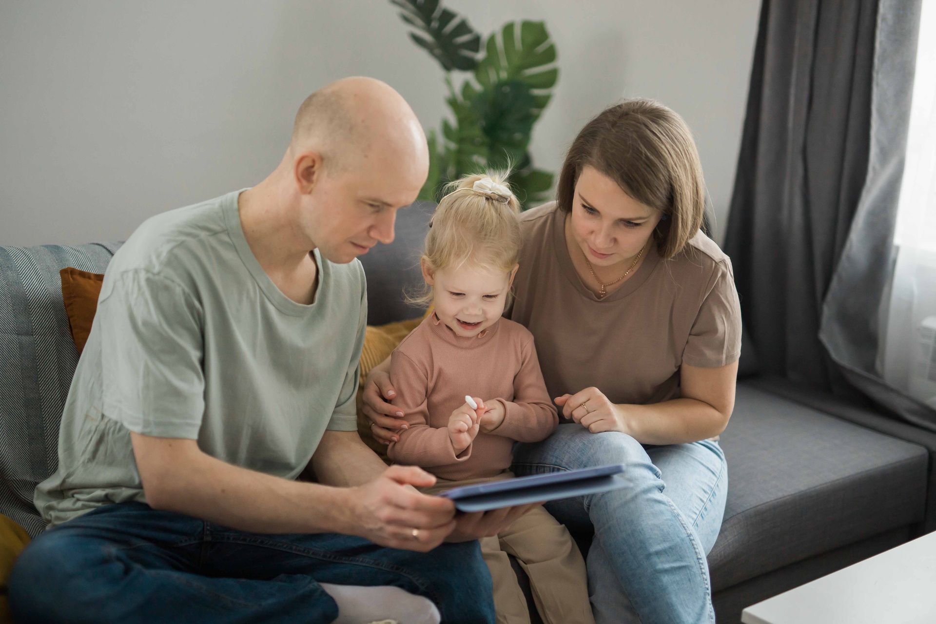 Family of three looks at tablet on couch. Girl smiles, parents look on.