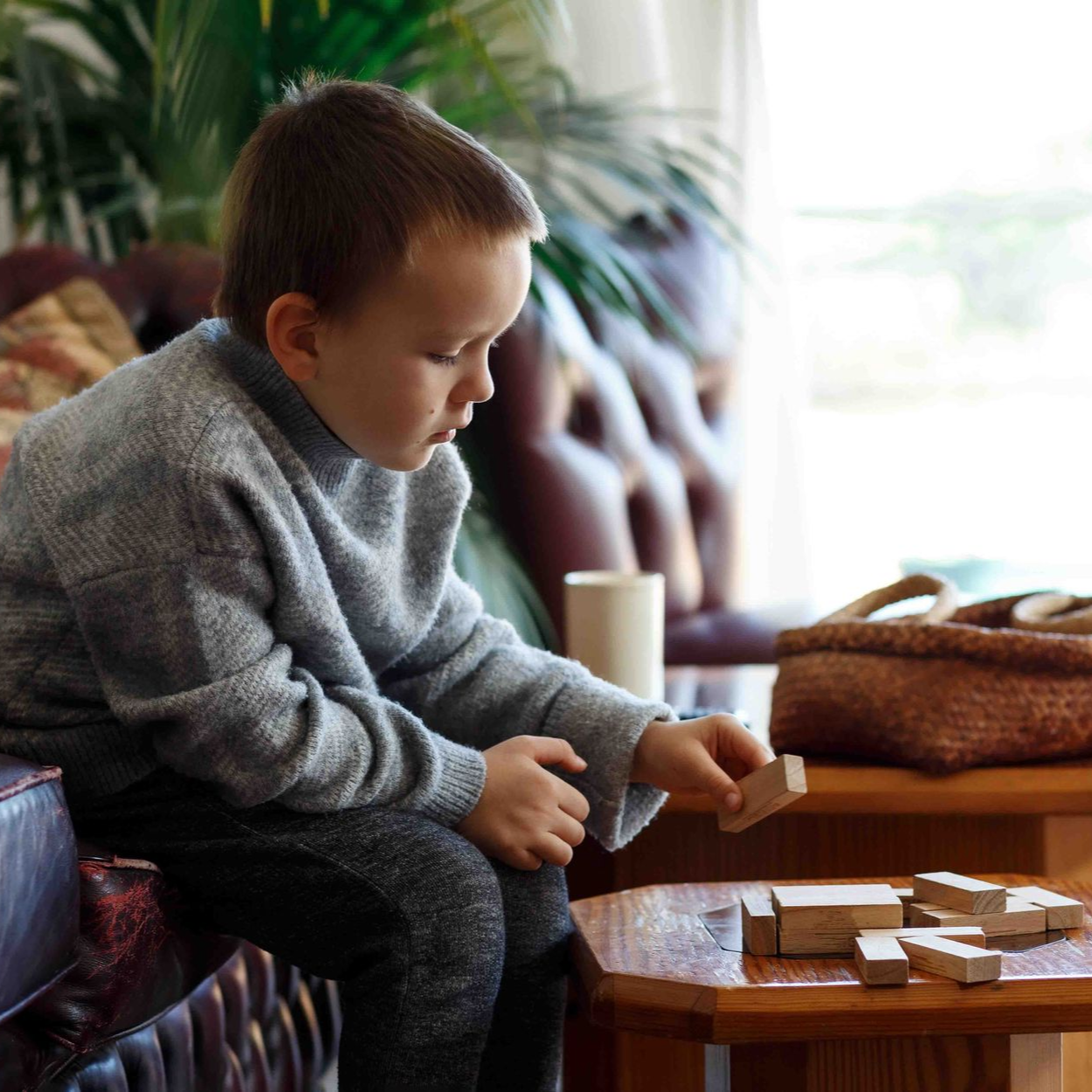 Boy in a gray sweater playing with wooden blocks on a table near a window.