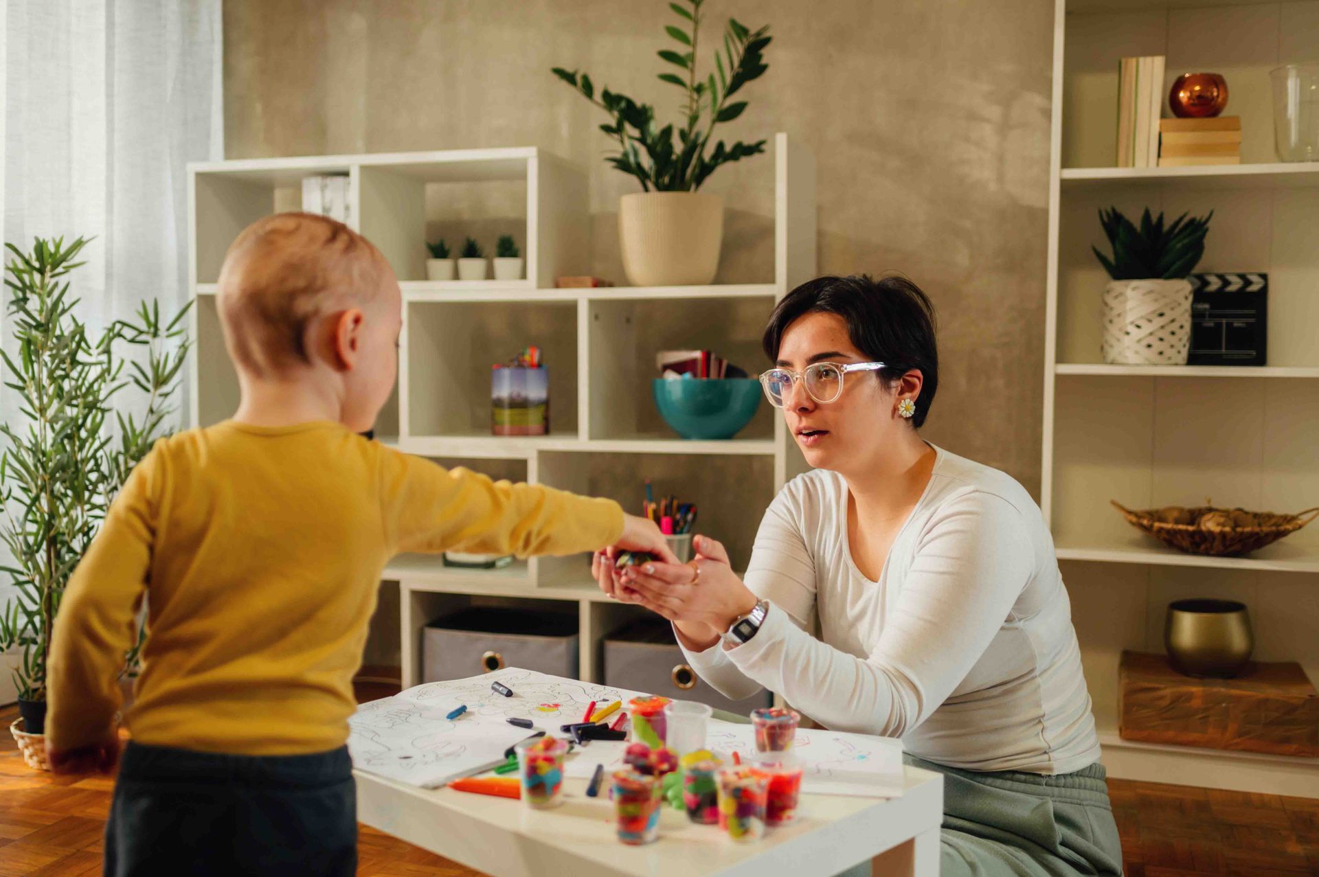 A person sits with a young child at a table with art supplies. They are interacting, possibly coloring.