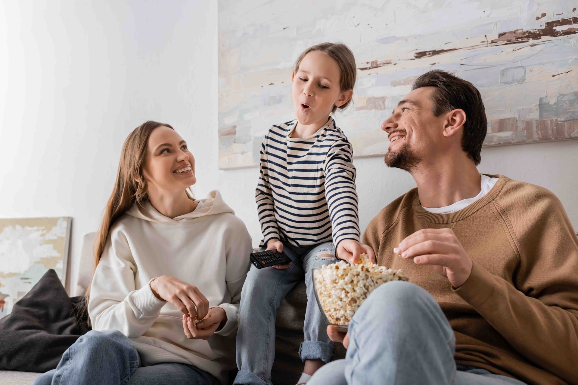 Family on a couch, child holding a controller, reaching for popcorn. Smiling, casual attire, indoor setting.