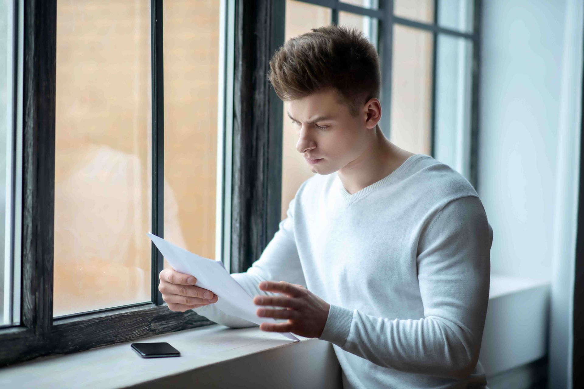 Man in white sweater reading papers by a window, looking concerned. Black phone on ledge.