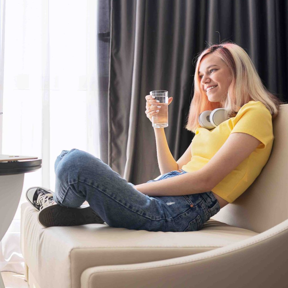 Young person in yellow top and jeans sits on a couch, holding a glass of water, smiling.