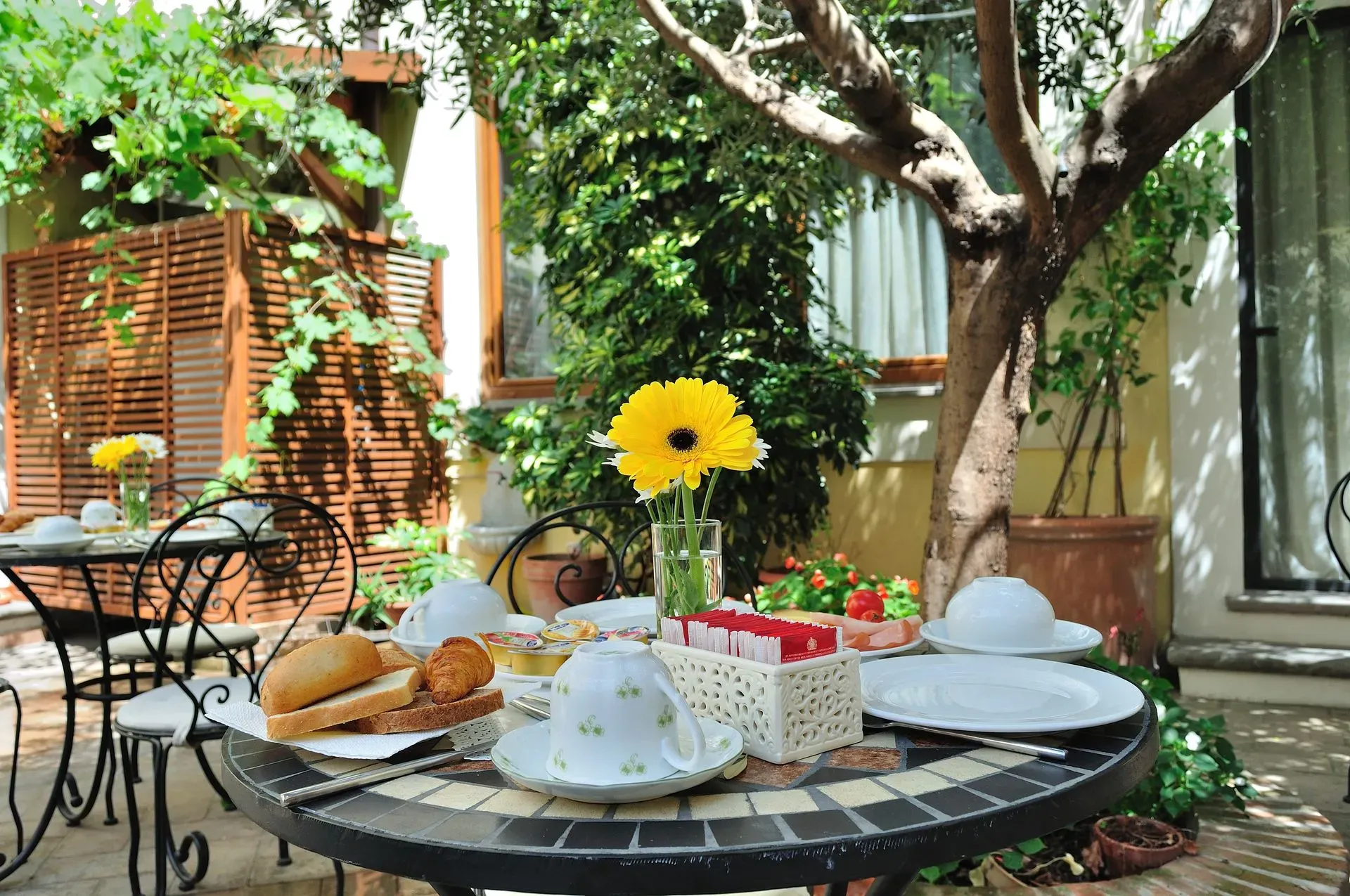 Tavolo per la colazione apparecchiato all'aperto, con pane, dolci e fiori. Ambiente soleggiato, da giardino.