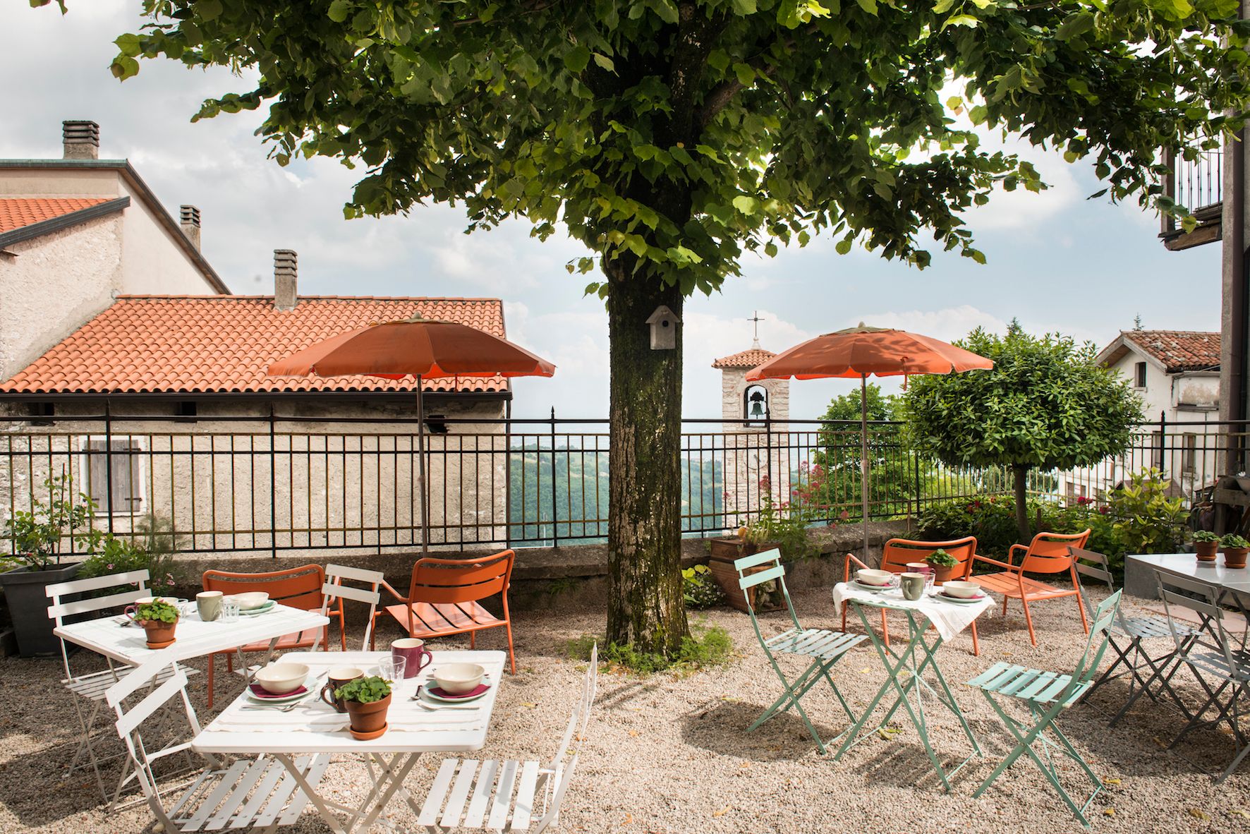 Locanda La Scuola con tavoli e sedie in metallo bianco e verde sotto un albero con ombrelloni arancioni, vista sui tetti di tegole rosse.