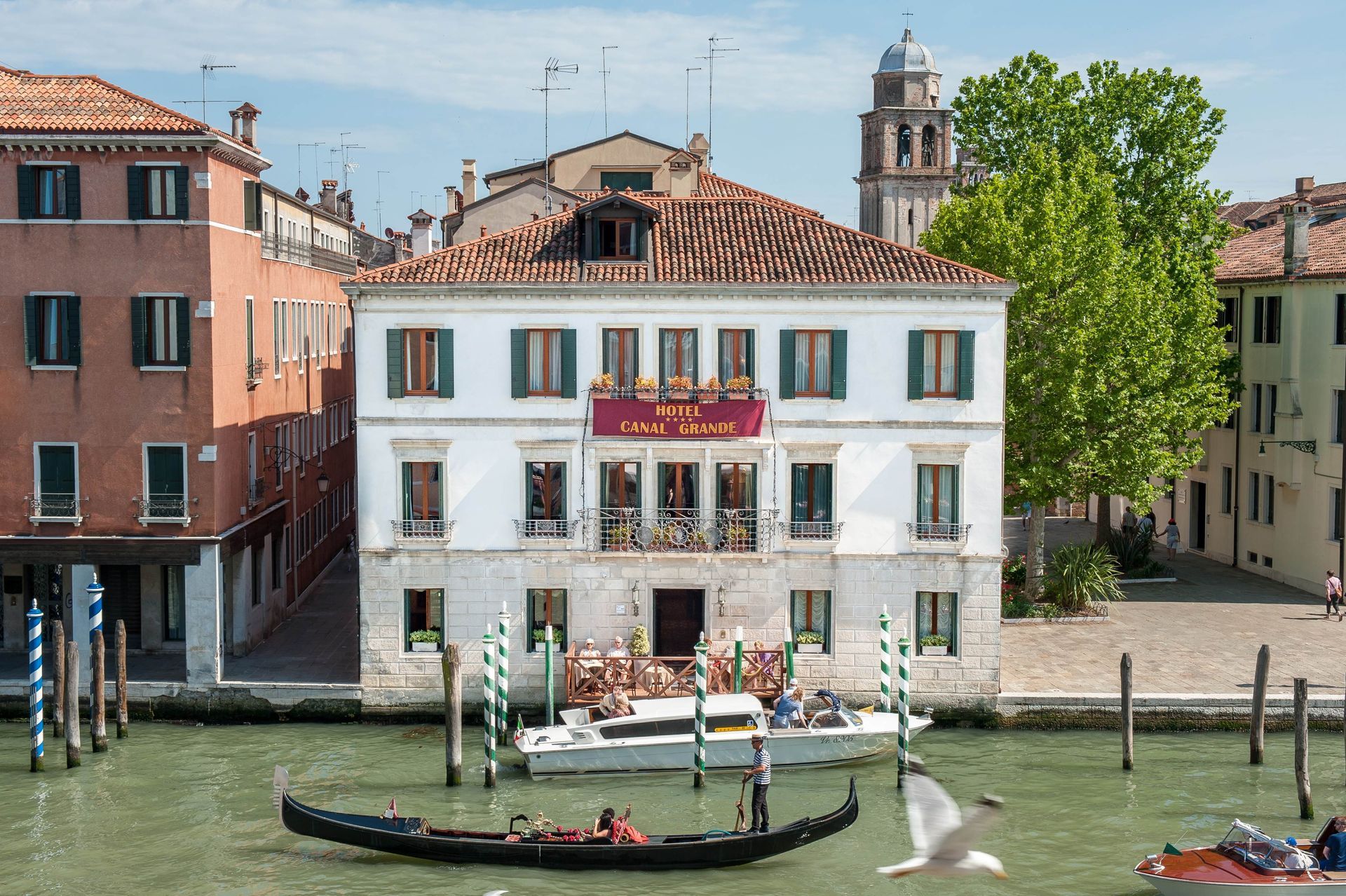 Scena di un canale veneziano con edifici, gondole, persone e una barca con musicisti.