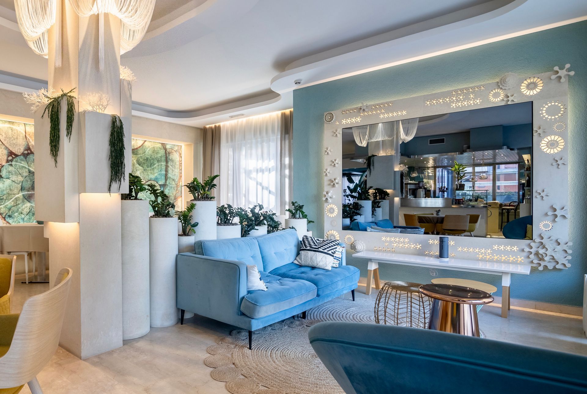 Divano azzurro in una stanza moderna e luminosa con grande specchio e accenti decorativi; piante in vaso.