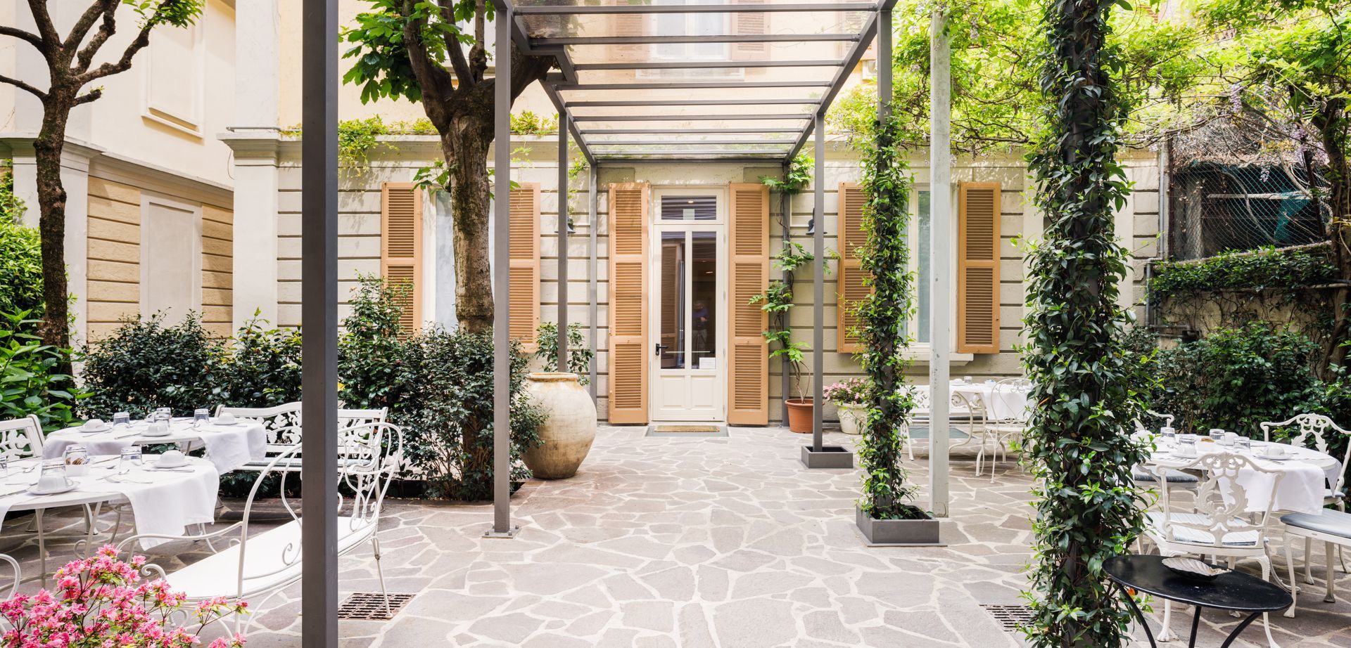 Patio esterno con tavoli bianchi, piante e un passaggio coperto. Edificio beige con persiane marroni aperte sullo sfondo.
