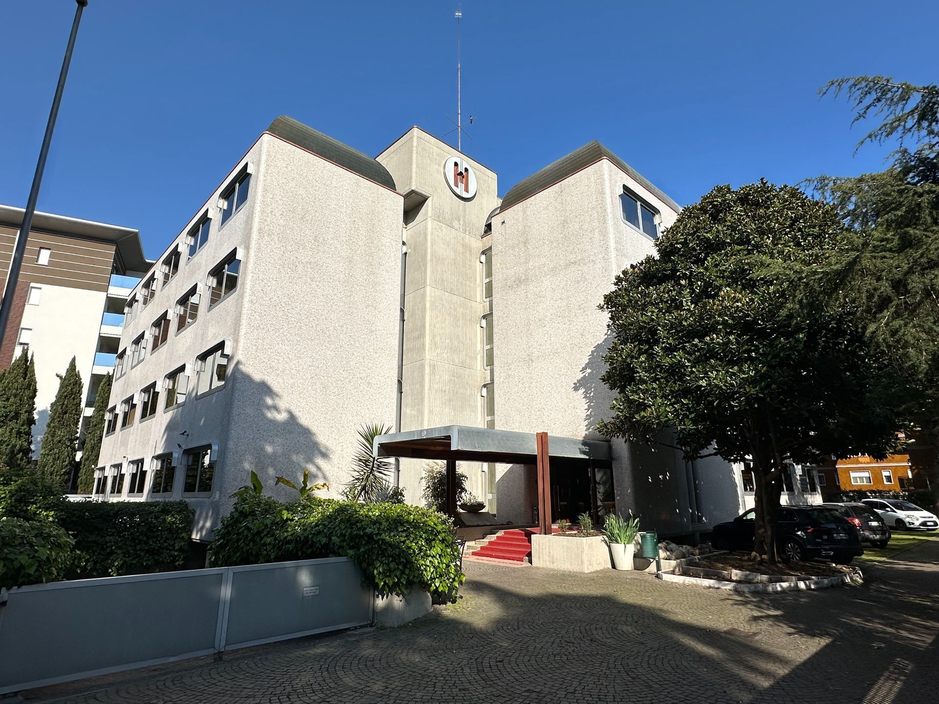 Un edificio grigio di quattro piani con il logo rosso sulla facciata, sotto un cielo azzurro.