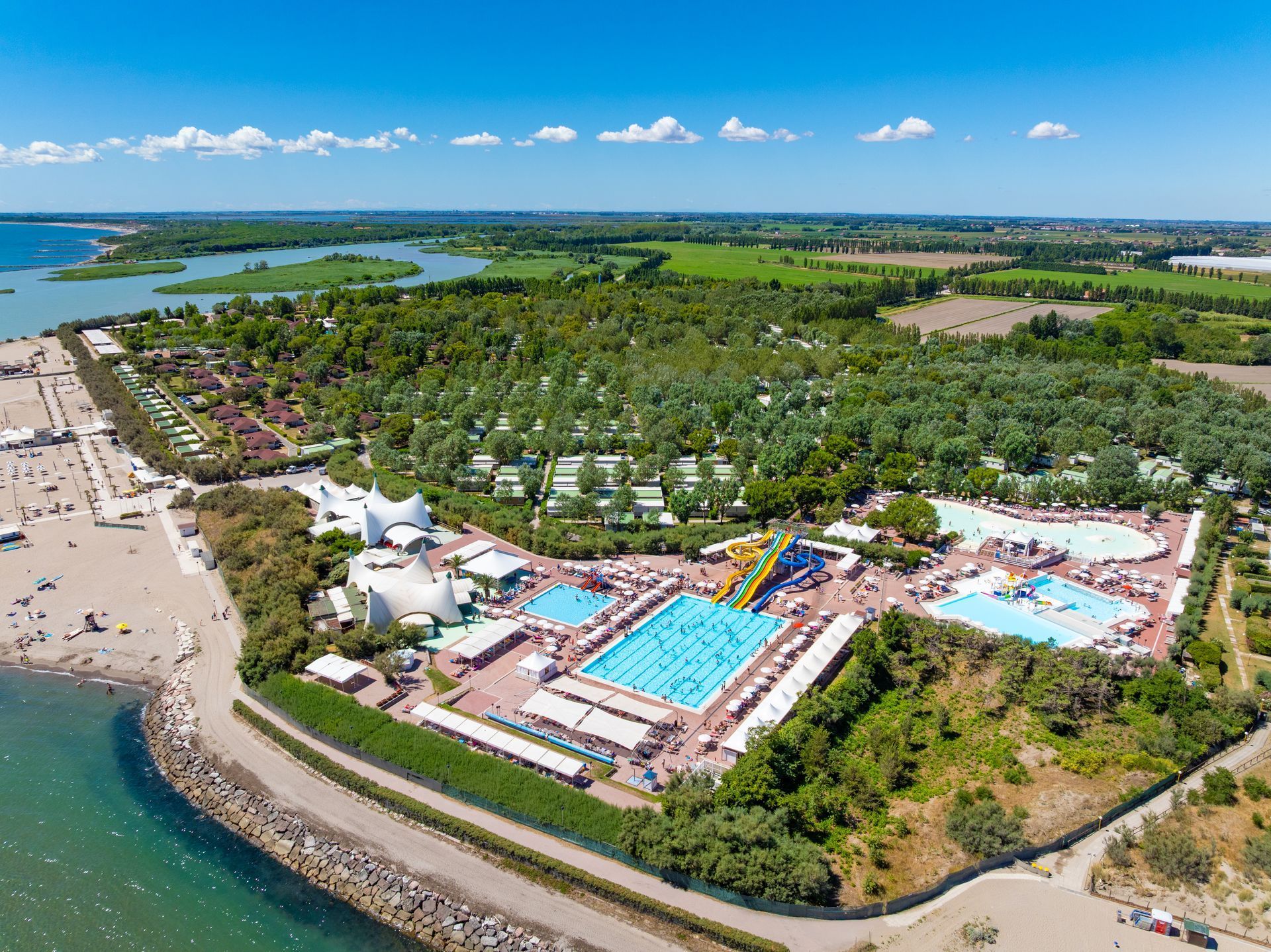 Vista aerea di un resort sulla spiaggia con piscine, alberi, edifici e una costa sotto un cielo azzurro brillante.