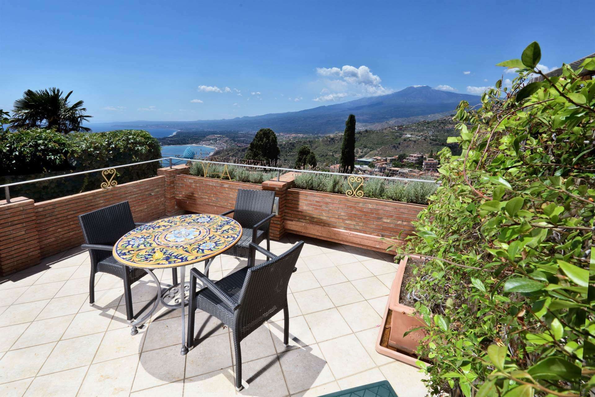 Patio con tavolo e sedie, con vista sul mare e sulle montagne in lontananza sotto il cielo azzurro.