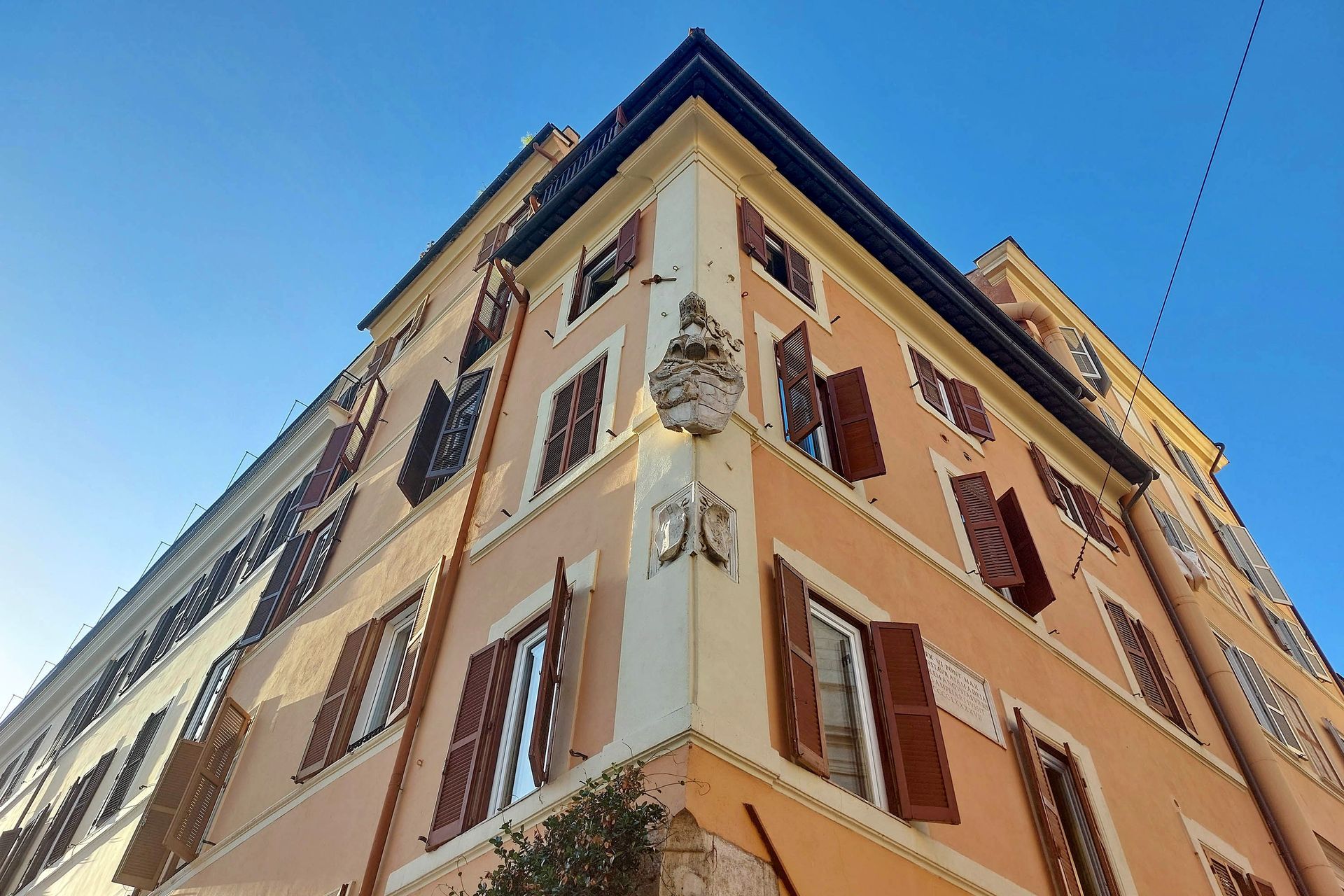 Vista dal basso dell'angolo di un edificio color pesca a Roma, con persiane marroni e un cielo azzurro brillante.