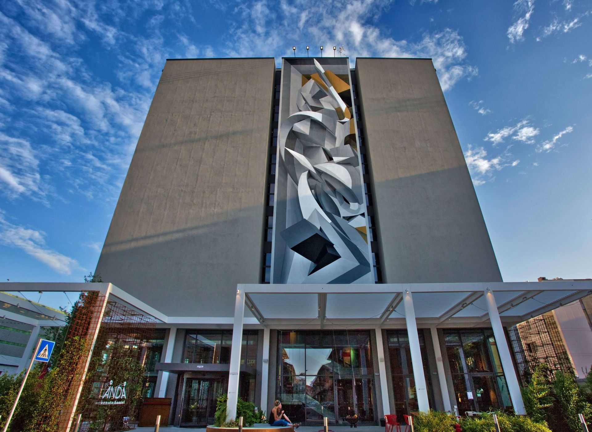 Alto edificio grigio con un grande murale astratto sulla facciata, sormontato da un cielo azzurro brillante.