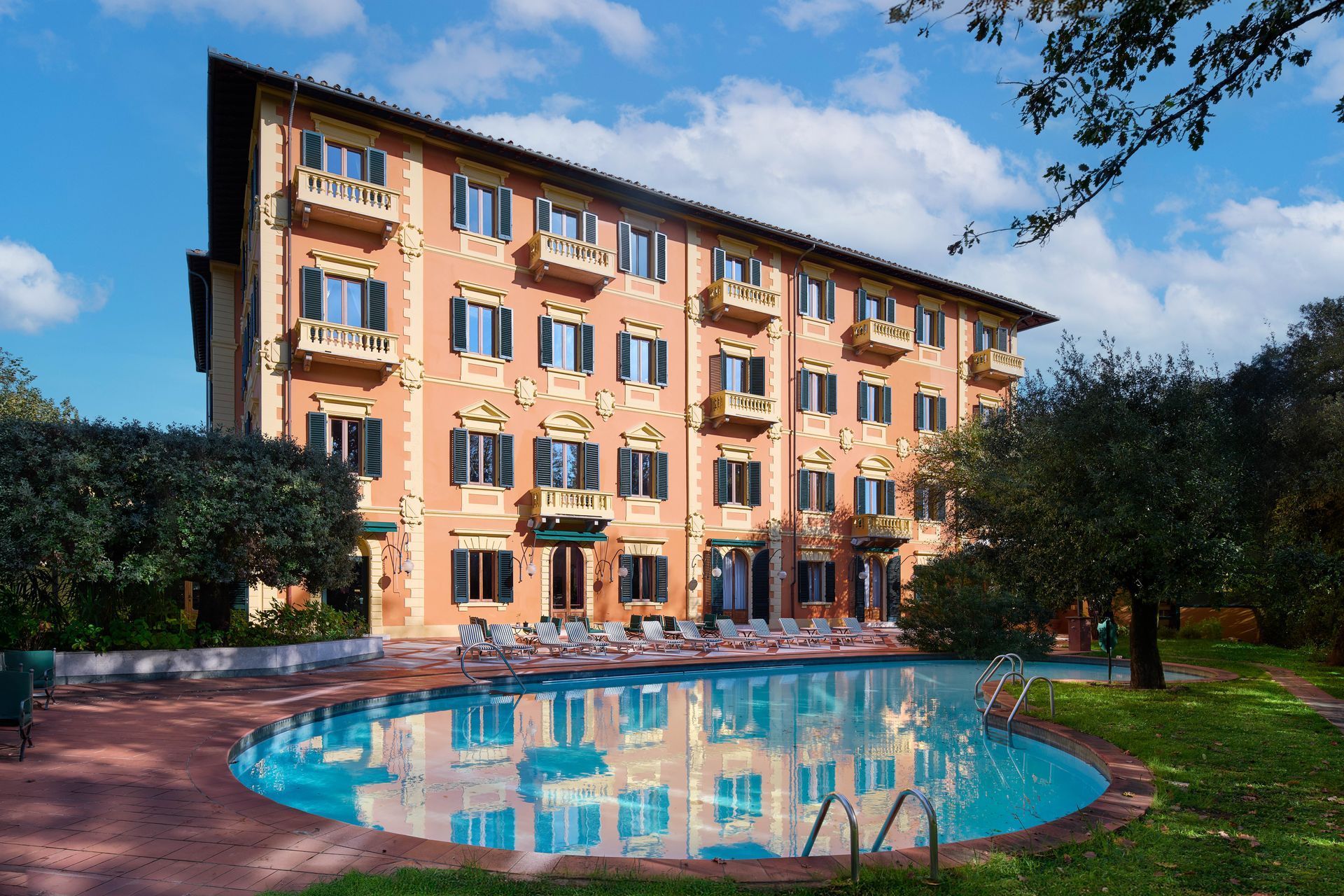 Hotel con piscina; edificio color salmone, finestre, balconi, piscina blu, sedie a sdraio, alberi, giornata di sole.